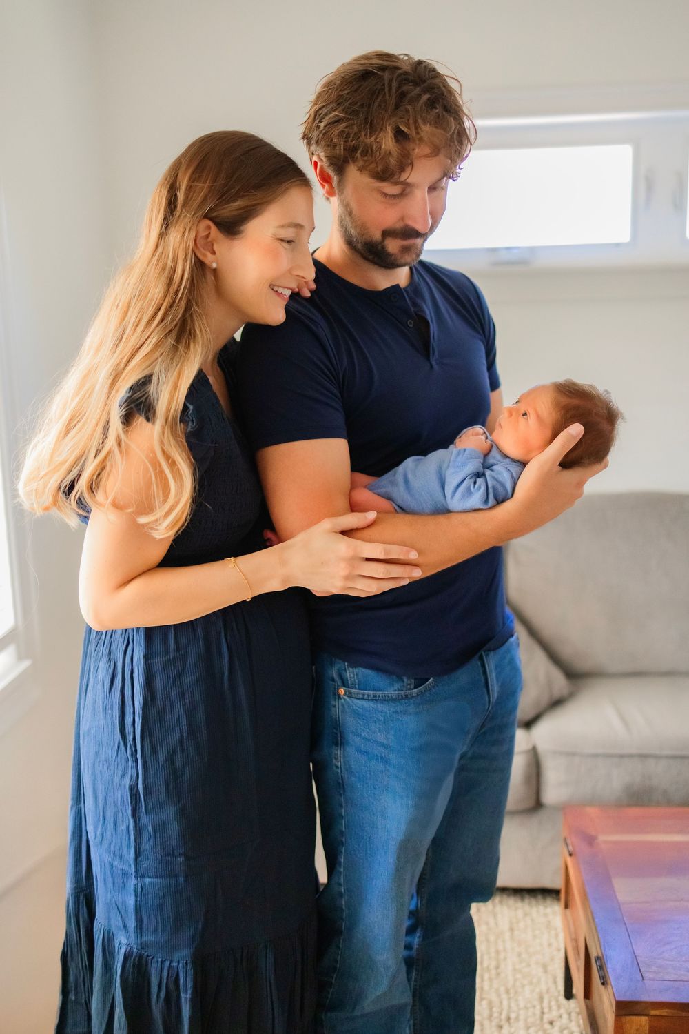 Young couple in navy blue outfits smiling at their newborn baby in a bright modern living room.