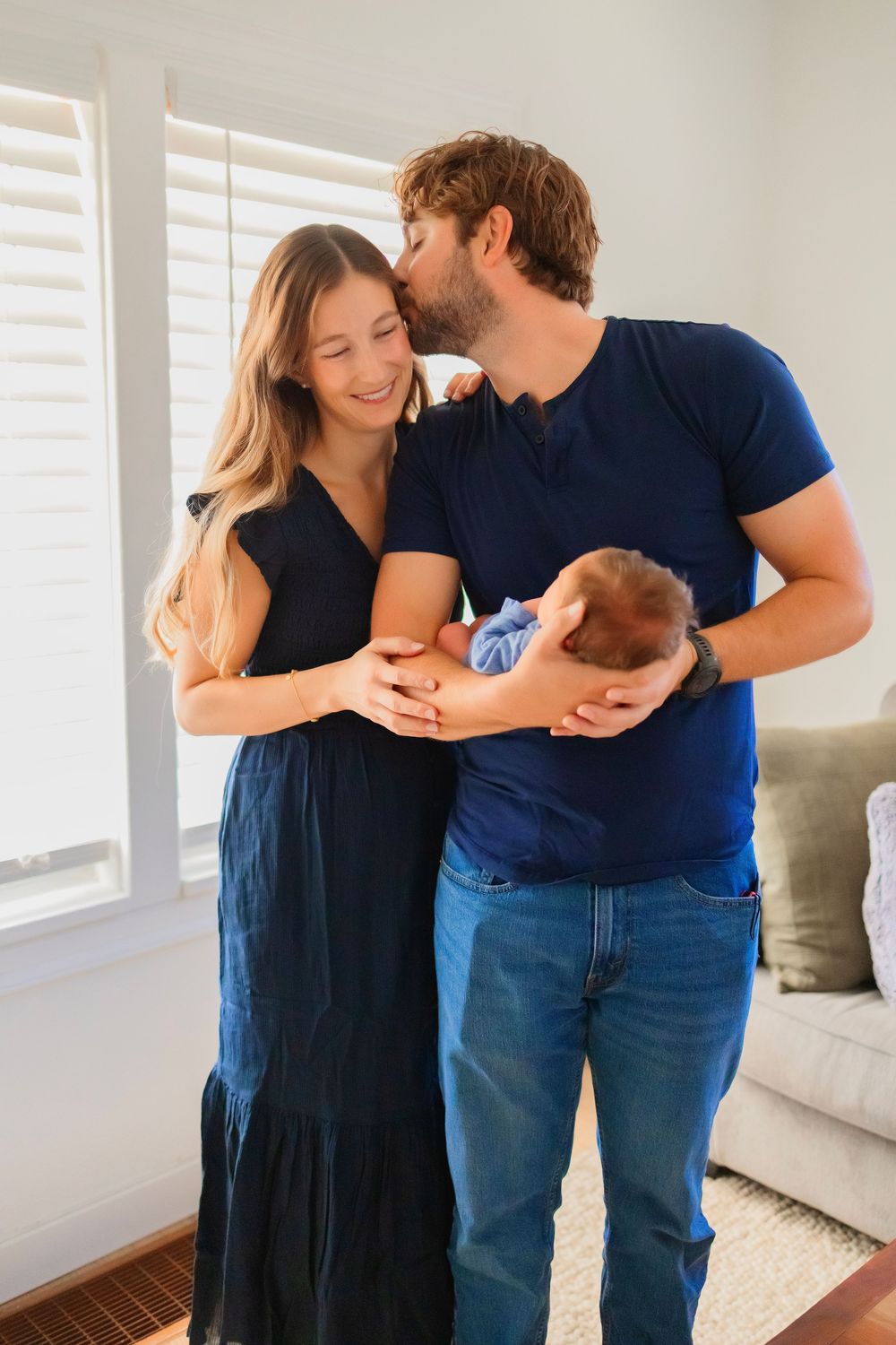 A couple in navy outfits smiles while cradling their newborn baby indoors near bright windows.