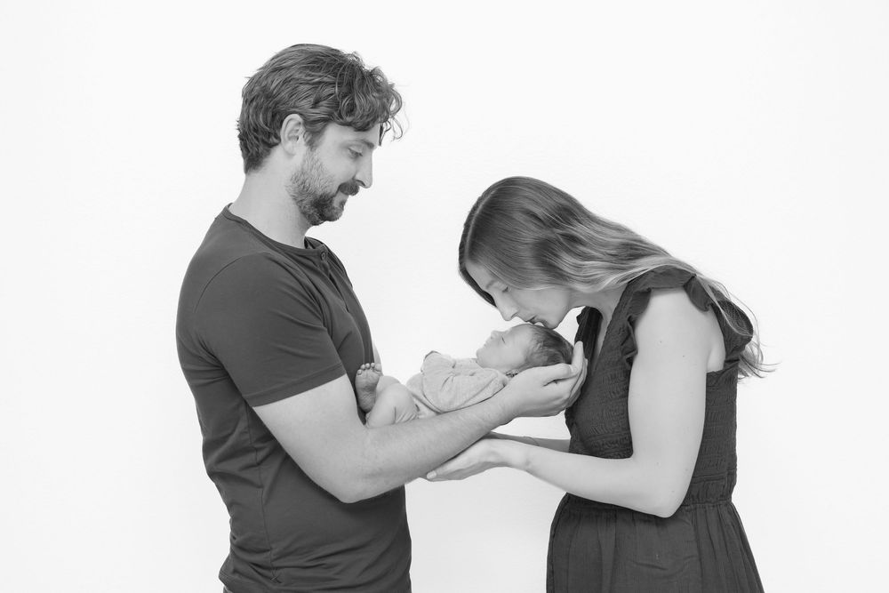 Black and white photo of parents tenderly holding and kissing their newborn baby against a white background.