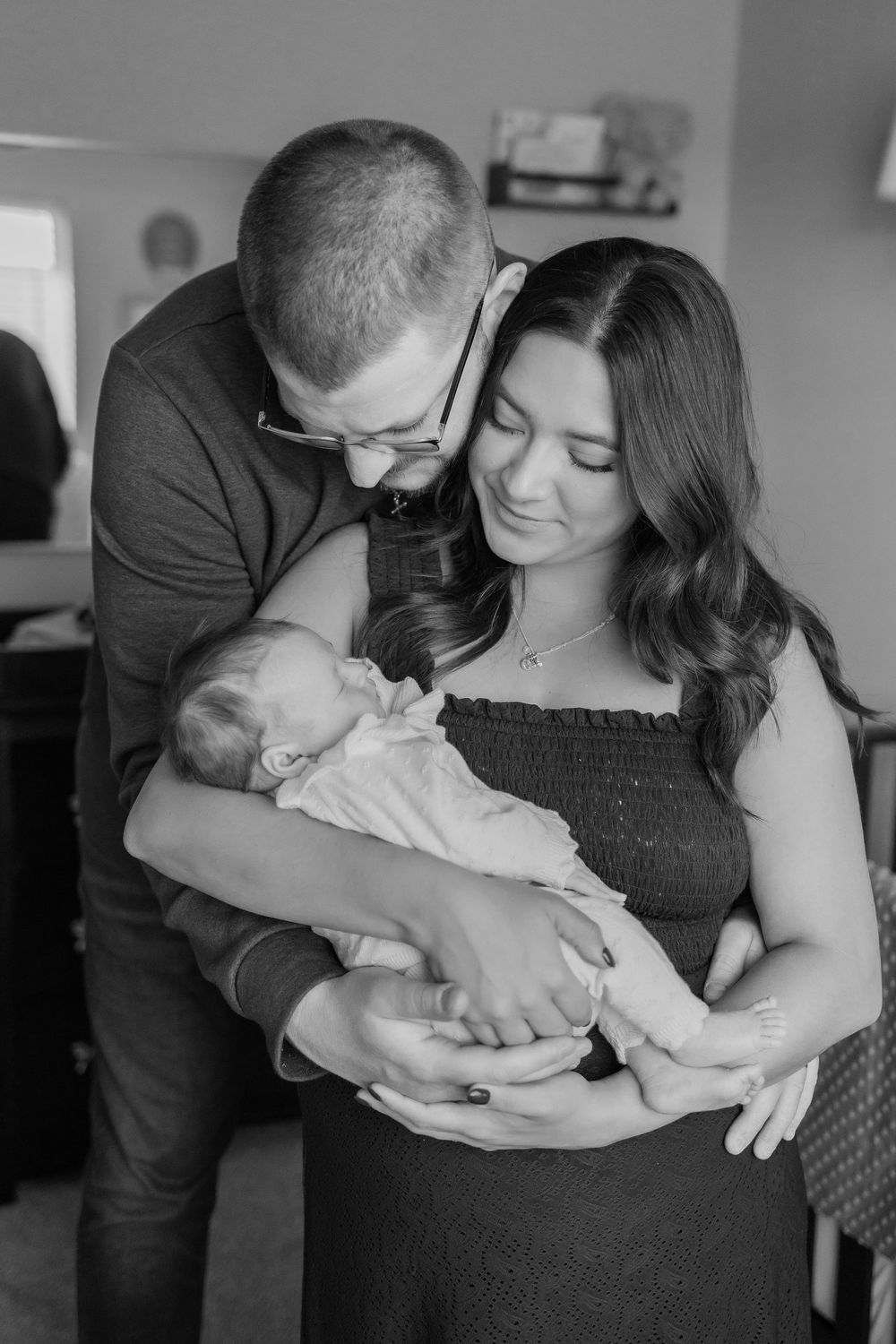 Black and white photo of new parents tenderly cradling their newborn baby together indoors.