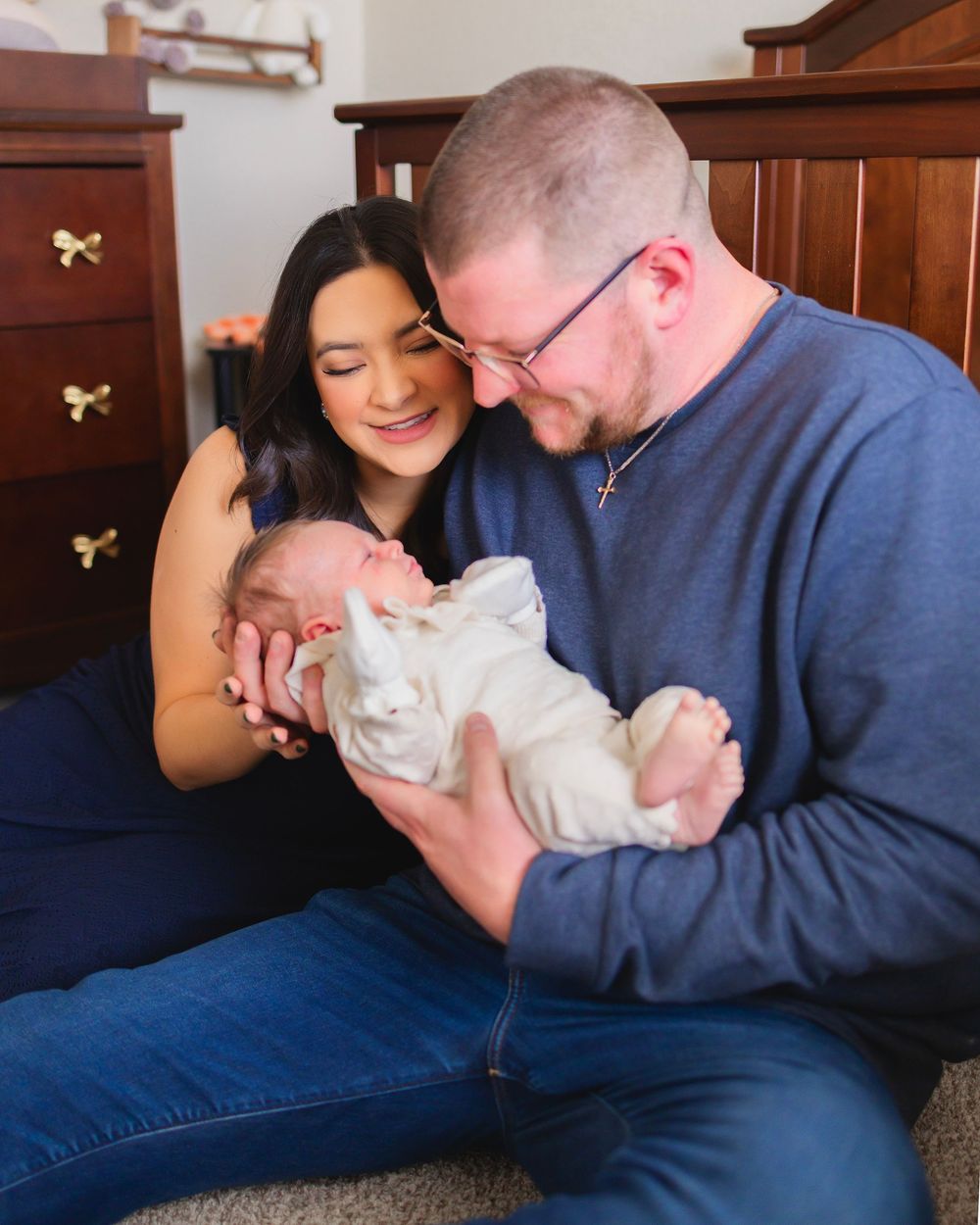 A couple sits on the floor smiling at their newborn baby dressed in white, with wooden furniture in the background.