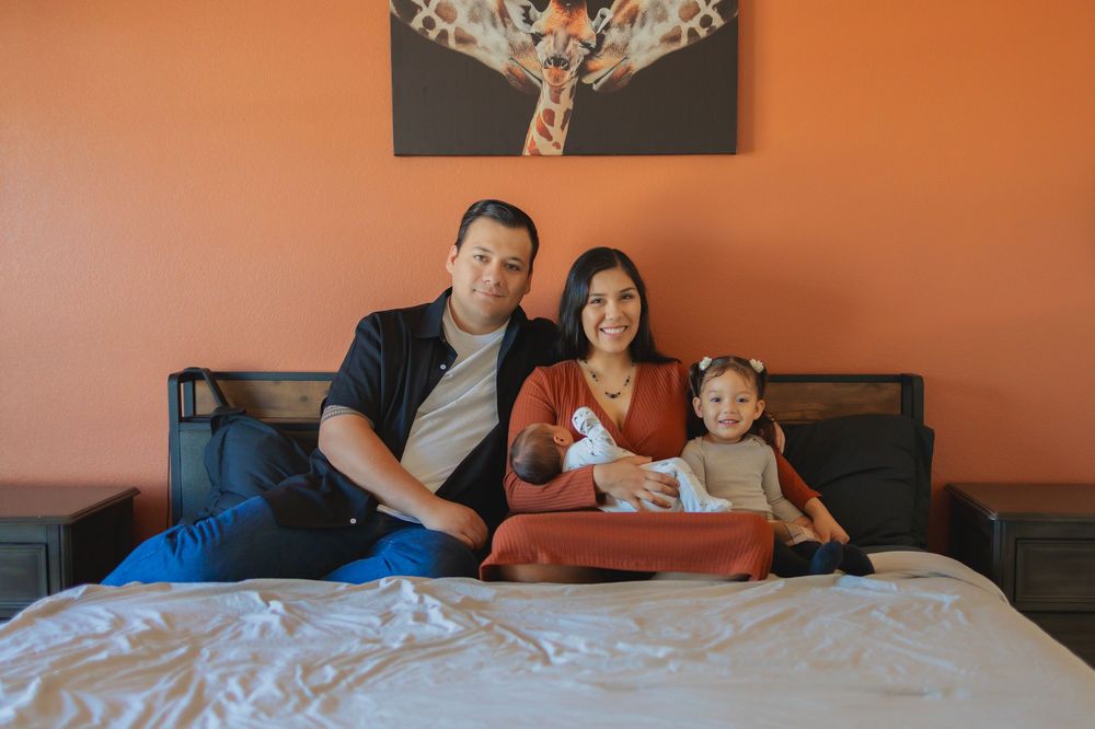 Happy family of four posing on a bed with orange walls and a giraffe painting, parents holding newborn baby.
