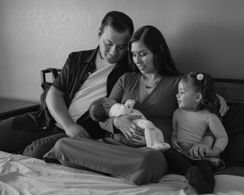 Black and white photo of a family with two parents sitting on a bed, holding a newborn baby, with a toddler beside them.