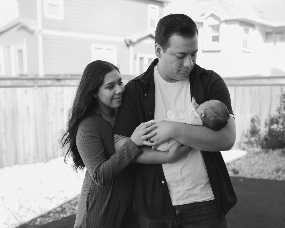 Black and white photo of a couple lovingly gazing at their newborn baby outdoors near a wooden fence.