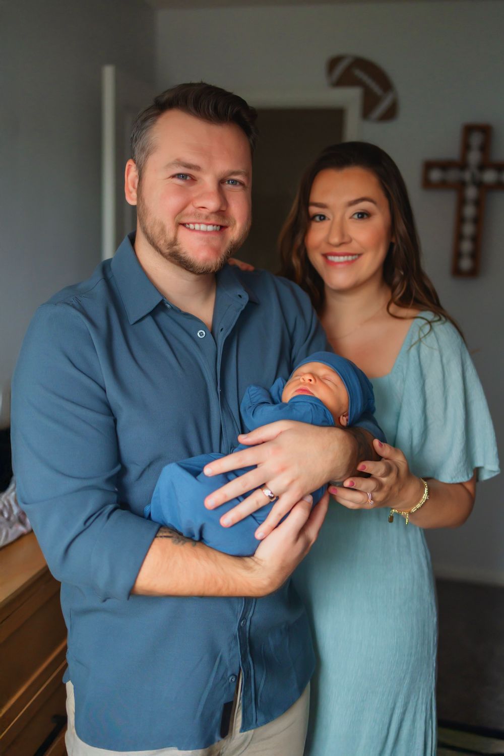 Happy couple smiling and holding their newborn baby dressed in blue inside a cozy home with a cross on the wall.