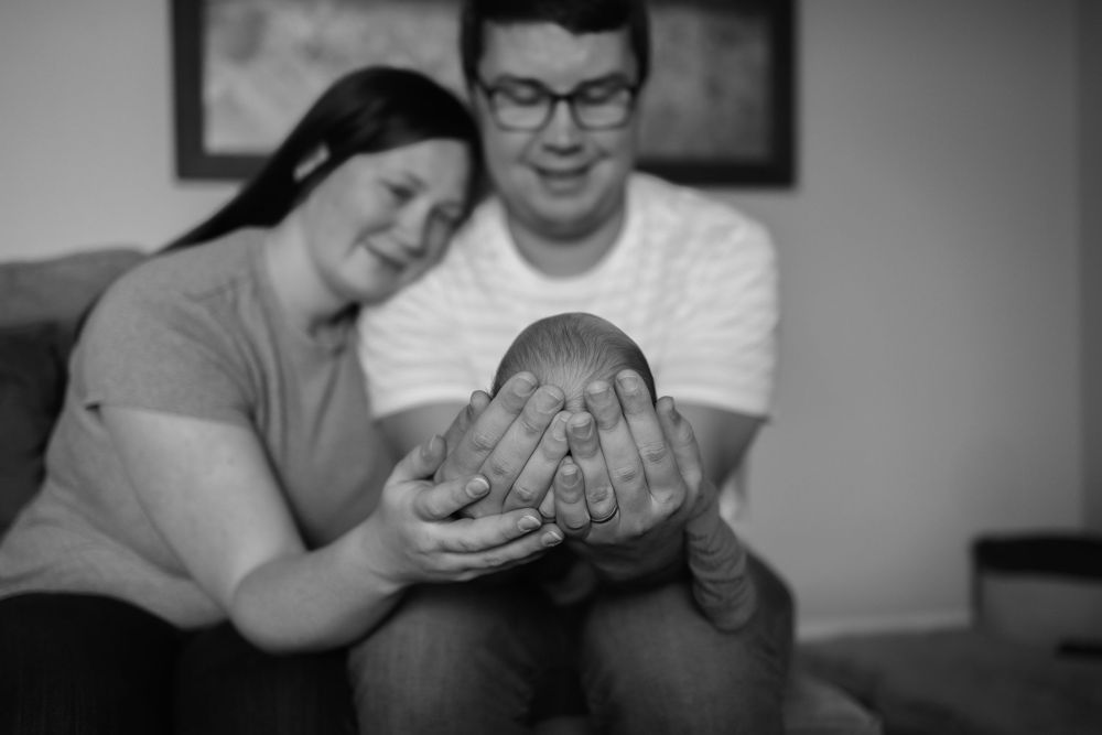 Black and white photo of smiling parents cradling their newborn baby together on a couch.