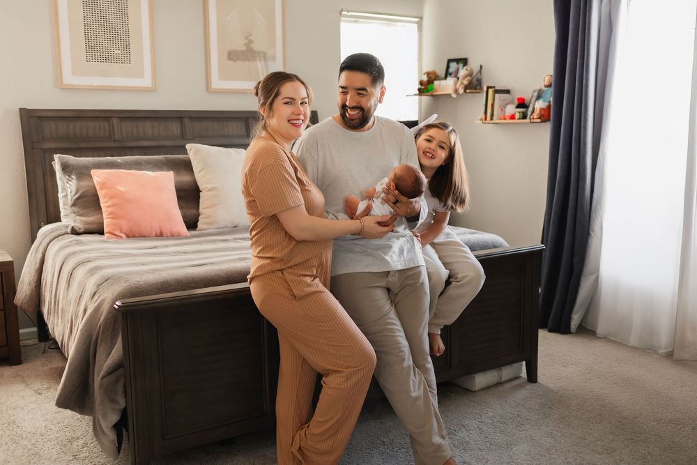 Happy family with newborn baby posing in a cozy bedroom, parents smiling and holding infant near a gray bed.
