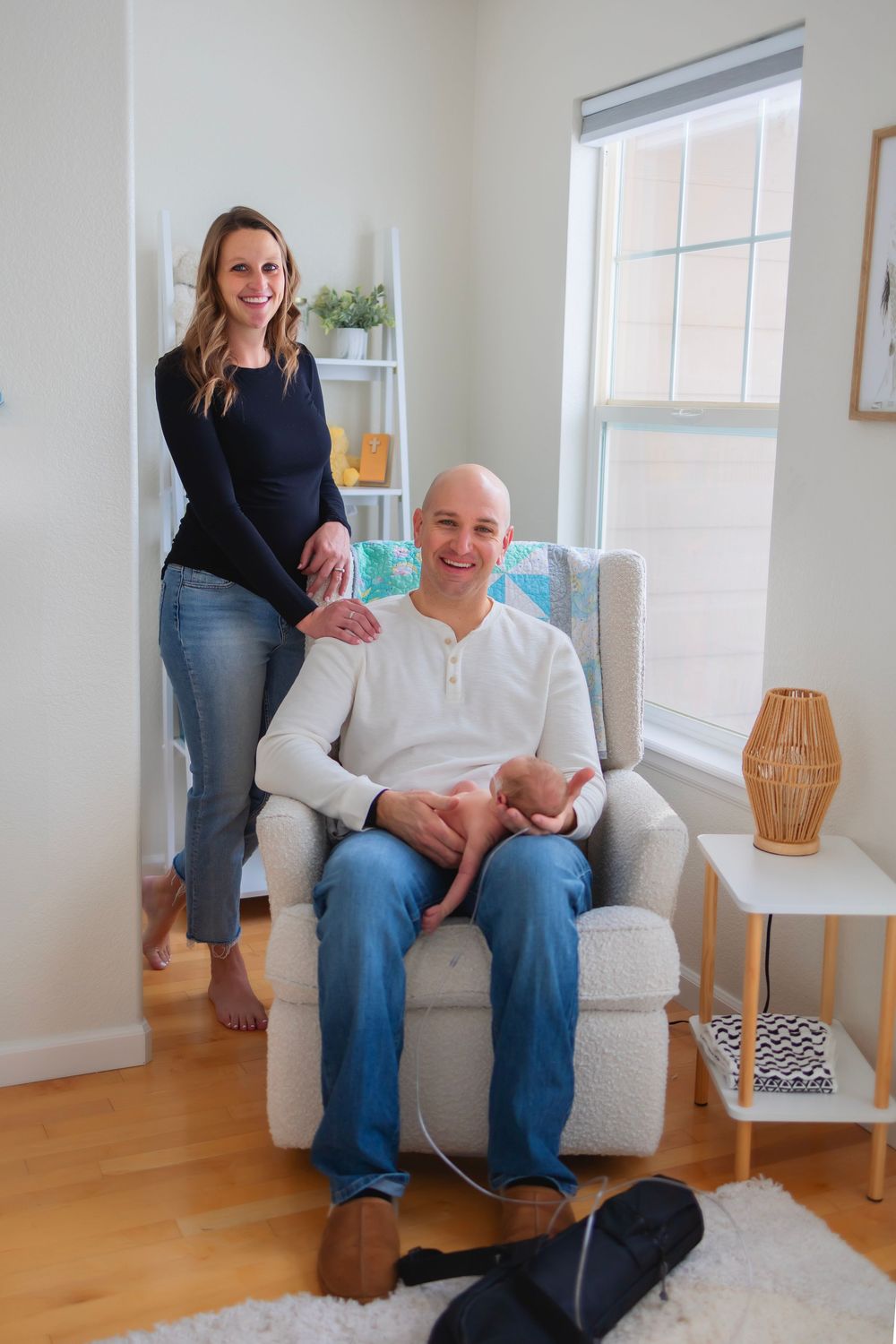A smiling couple poses in a cozy nursery, man holding newborn in armchair, woman standing beside him near a bright window.