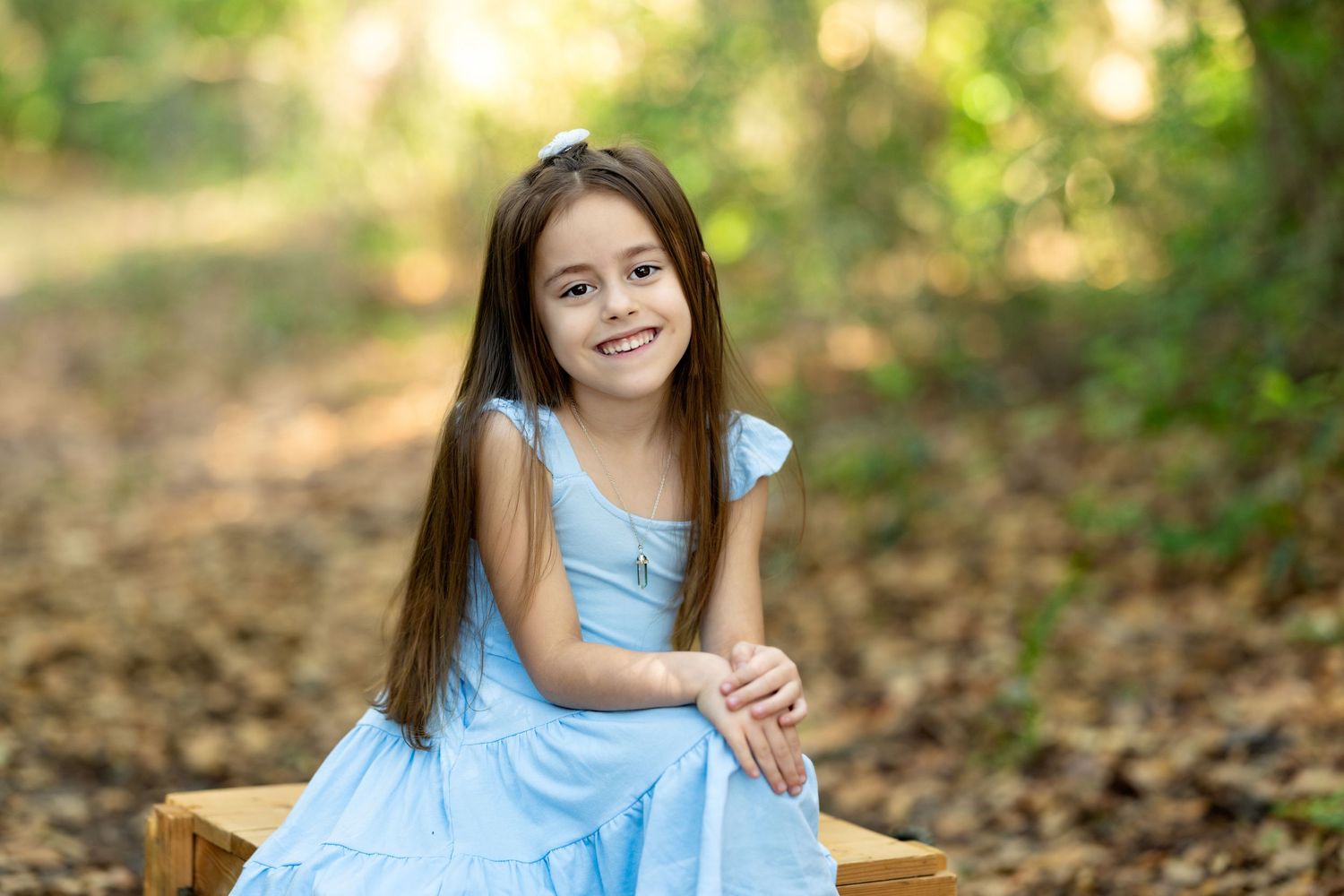 A smiling young girl in a light blue dress sits on a wooden bench surrounded by a lush green forest backdrop.