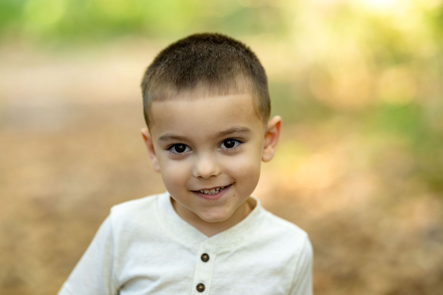 Smiling young boy with short hair wearing a white henley shirt outdoors in a natural green setting.