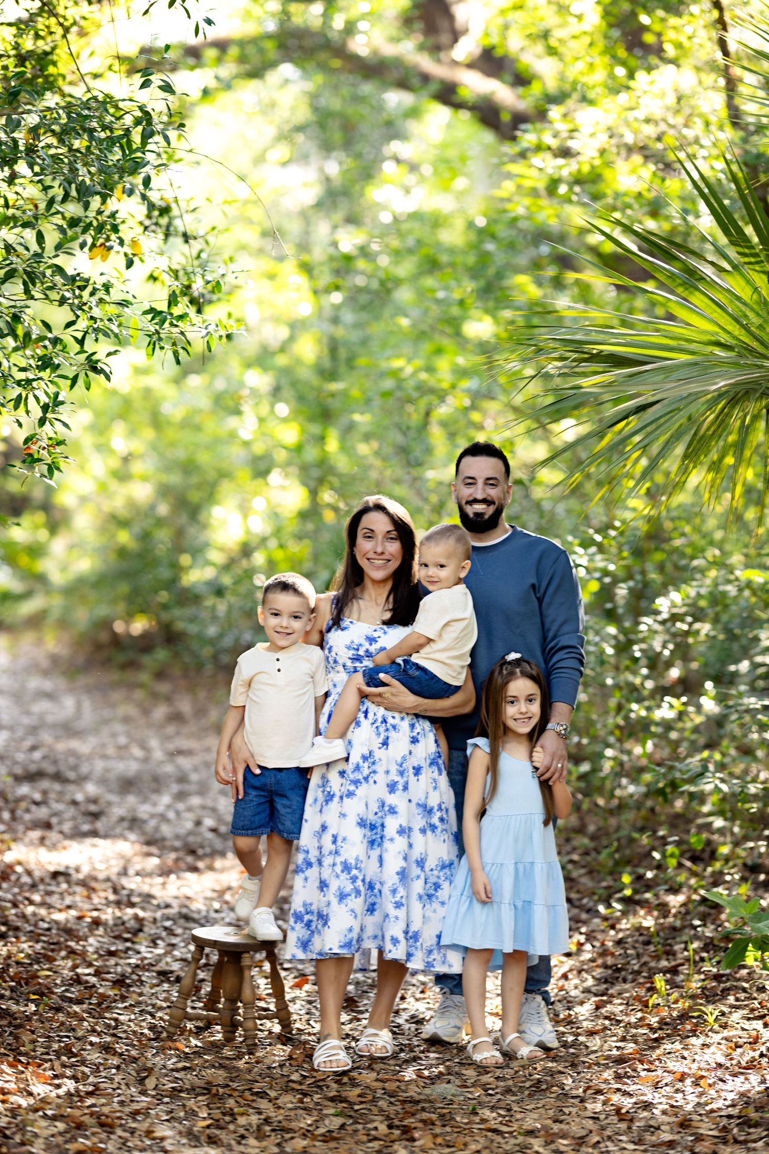 Family of five posing on a leaf-covered path surrounded by lush green trees in a sunlit outdoor setting.