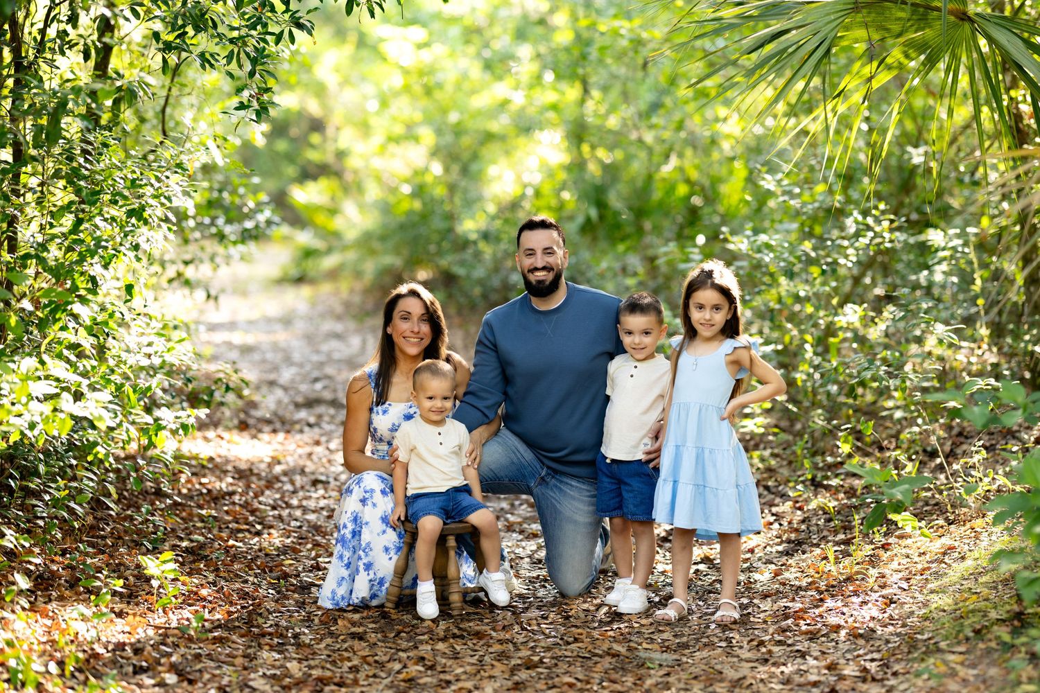 Family of five posing on a sunlit forest path surrounded by lush green trees and foliage.