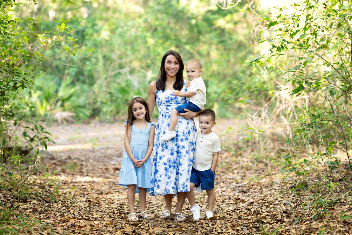 A mother in a floral dress poses with her three young children on a leaf-covered path surrounded by lush green trees.