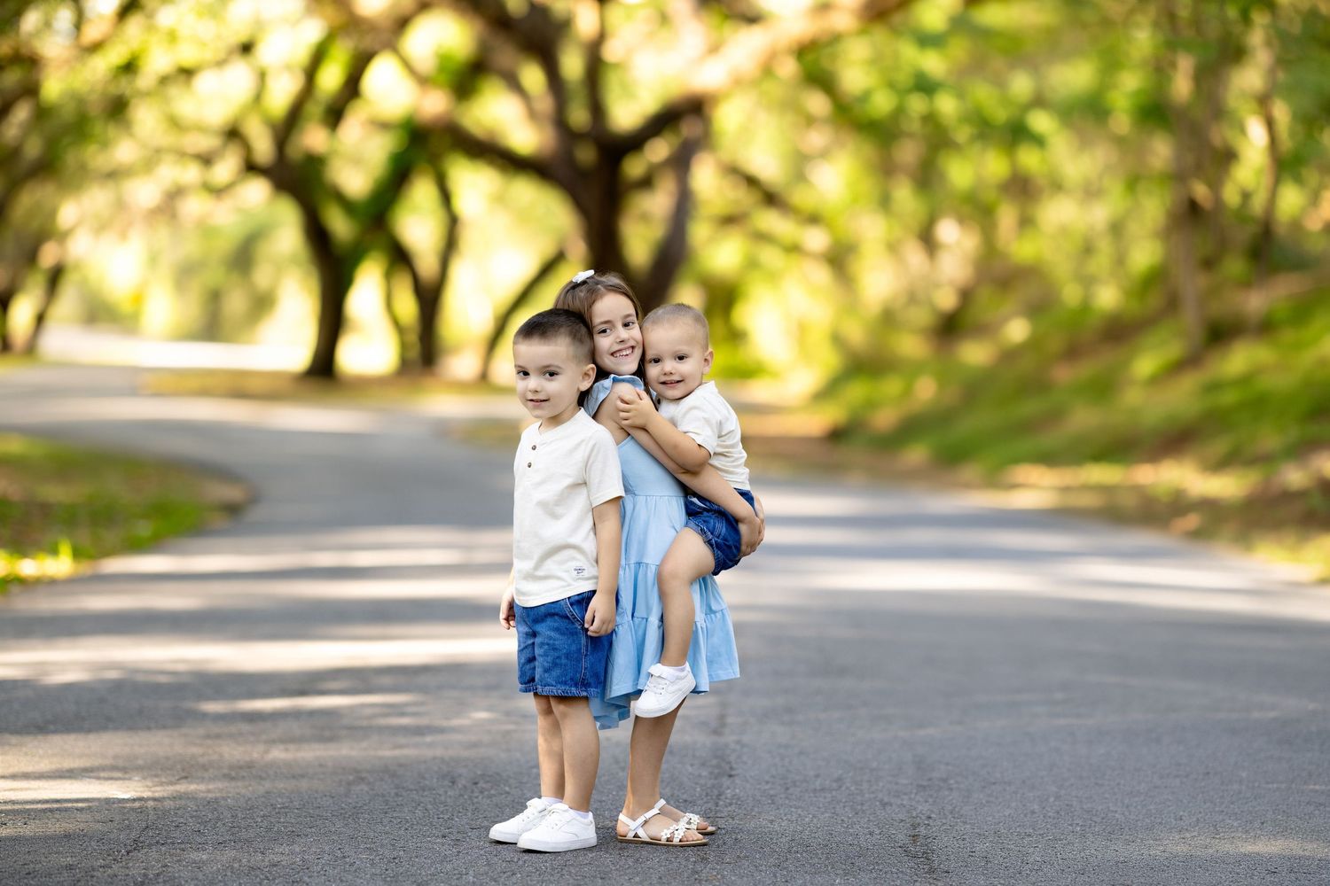 Three young siblings pose on a tree-lined road, with older girl holding a baby and boy standing in front.