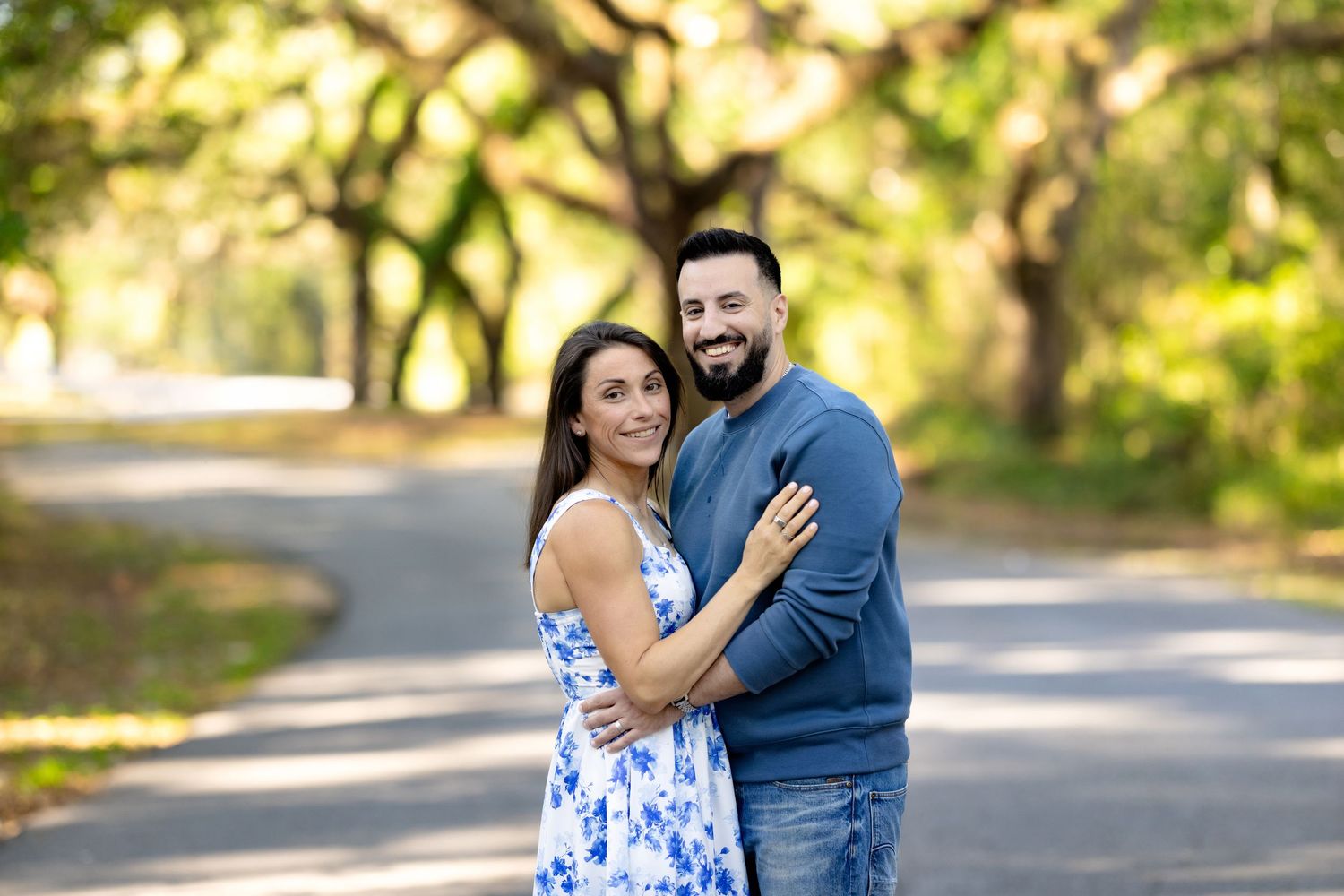 Couple smiling and embracing on a tree-lined road, woman in floral dress, man in blue sweater and jeans.