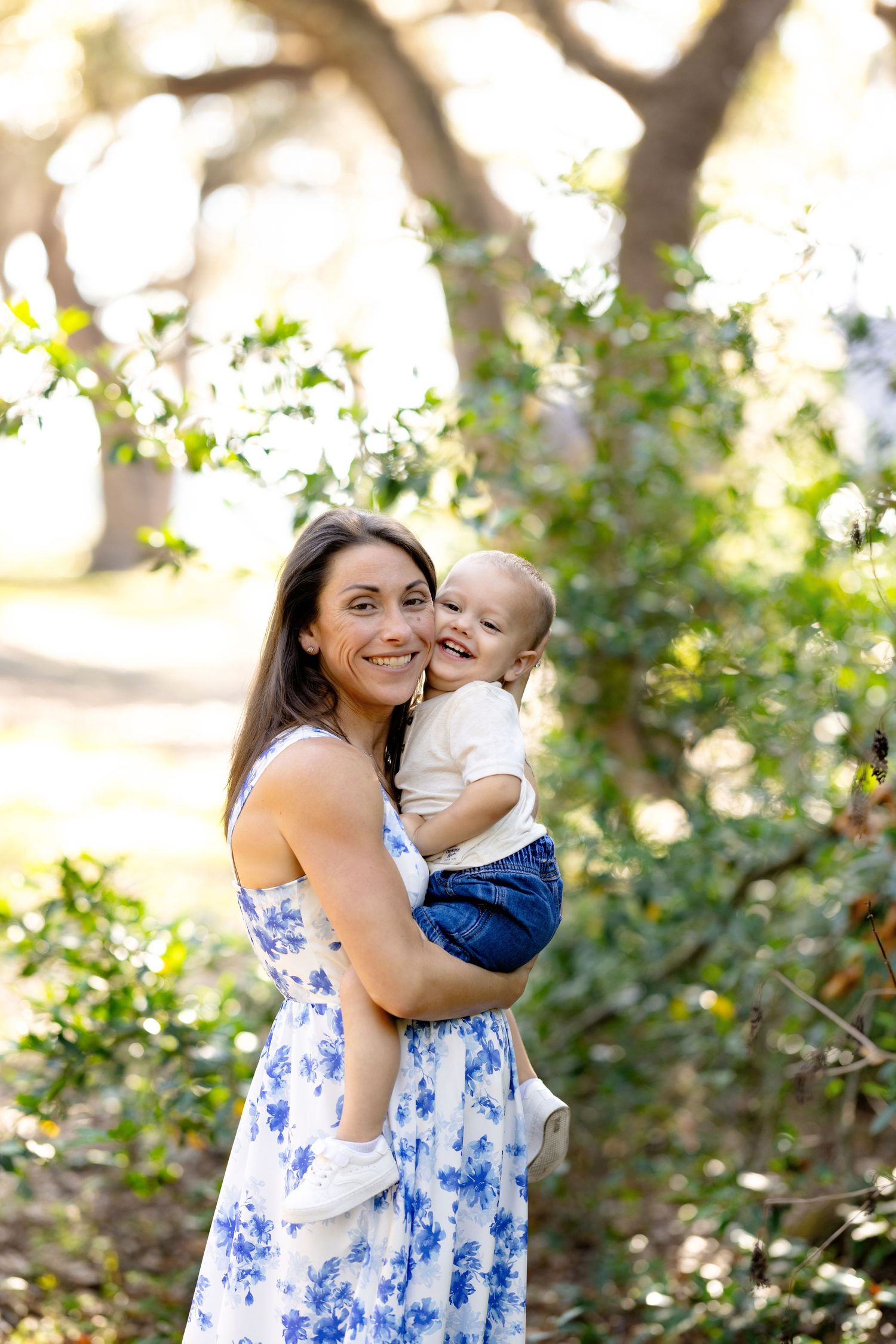Smiling woman in blue floral dress holding a happy baby outdoors in a sunlit park with green trees.