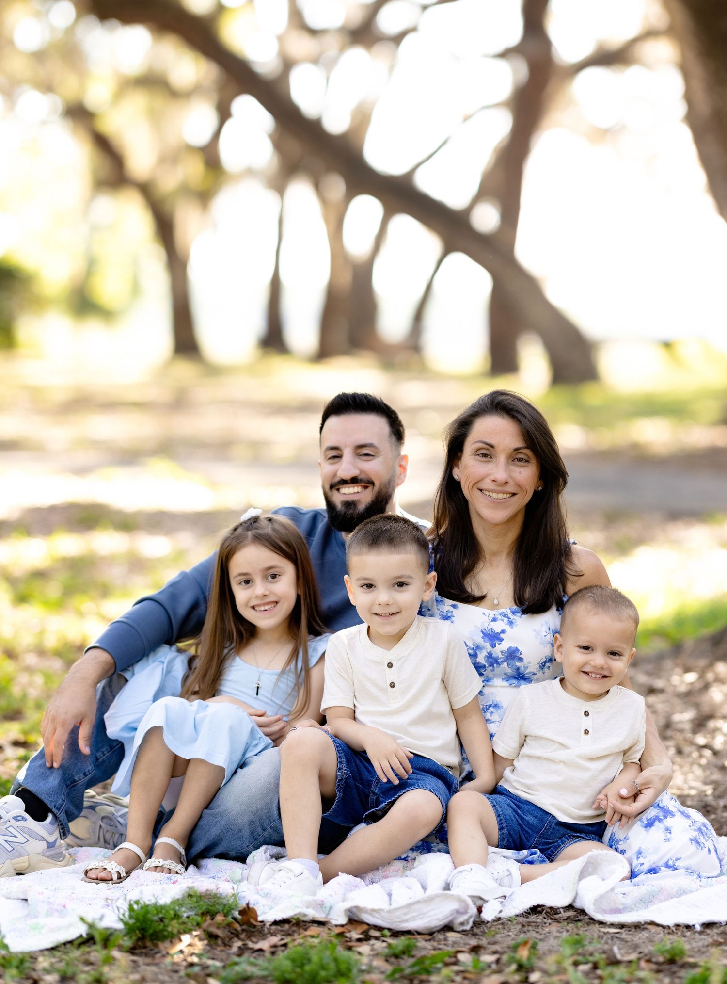 Family of five smiling together outdoors on a blanket in a sunlit park with trees in the background.