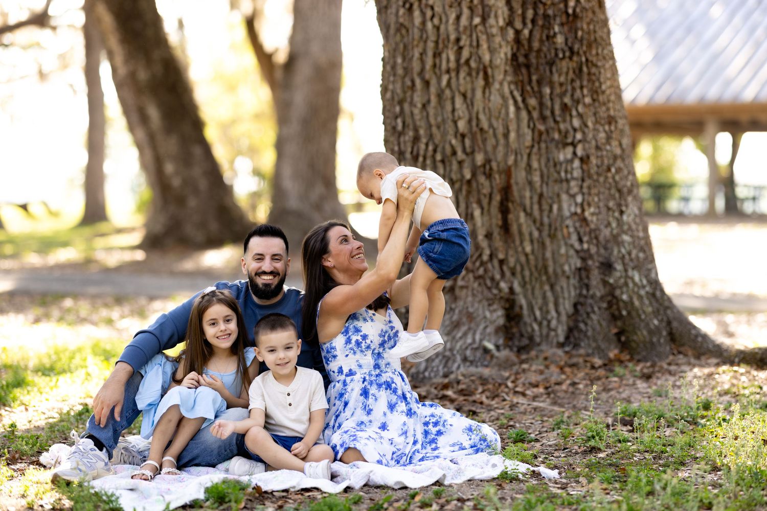 Family of five sitting on a blanket under large trees in a park, mother lifting baby while children smile.