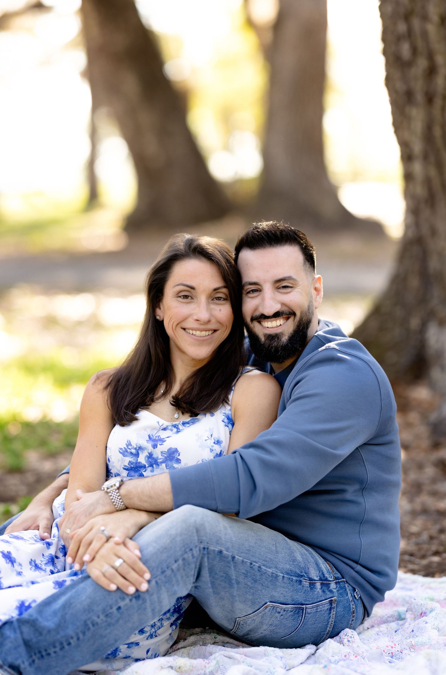 A smiling couple sits together outdoors in a park, woman in floral dress and man in blue hoodie, embracing warmly.
