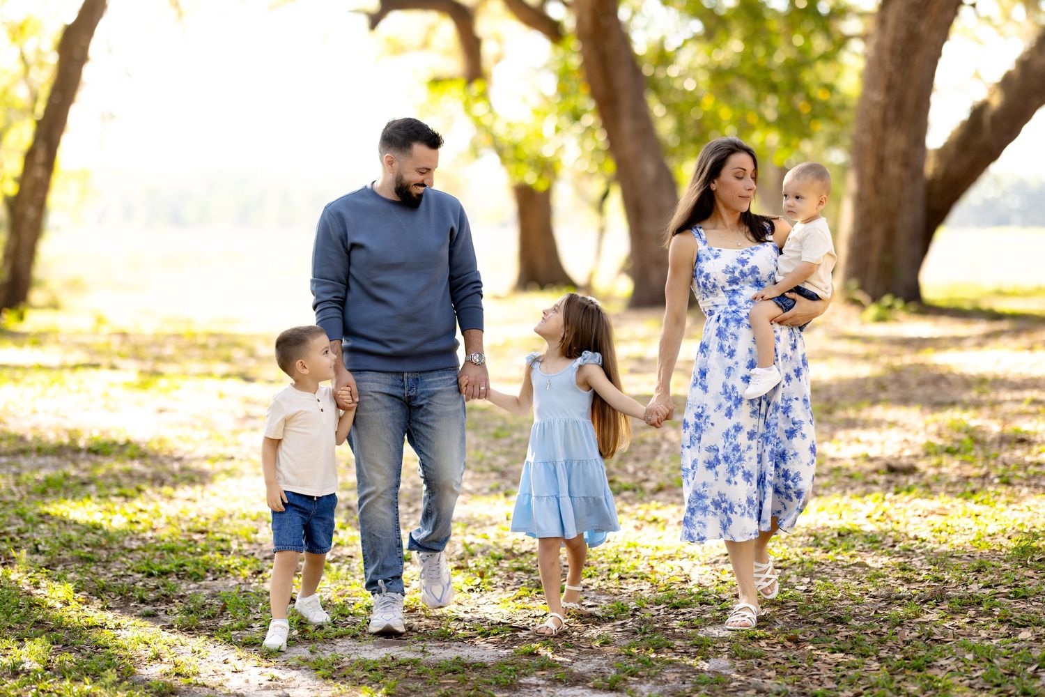 Family of five walking hand-in-hand through a sunny park, parents with three young children enjoying outdoor time together.