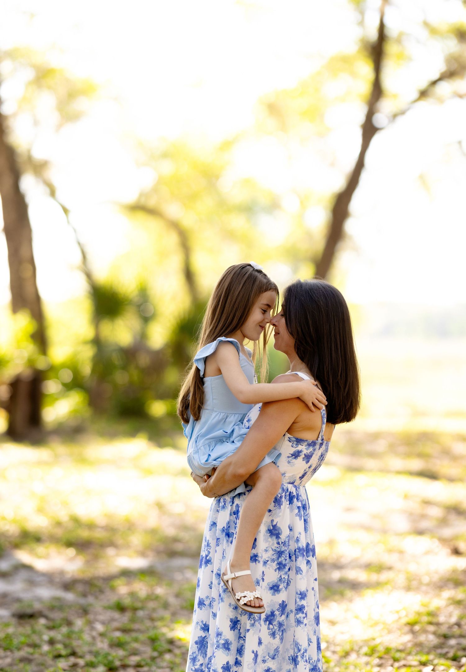 A mother holds her young daughter in a sunlit park, sharing a tender nose-to-nose moment outdoors.