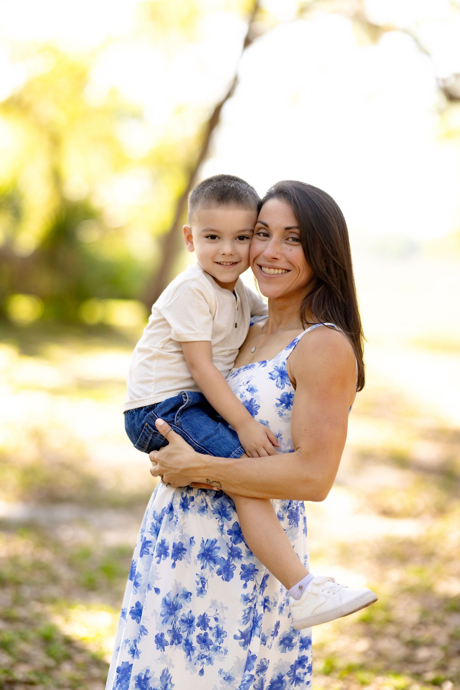 A smiling mother in a floral dress holds her young son outdoors in a sunlit park setting.