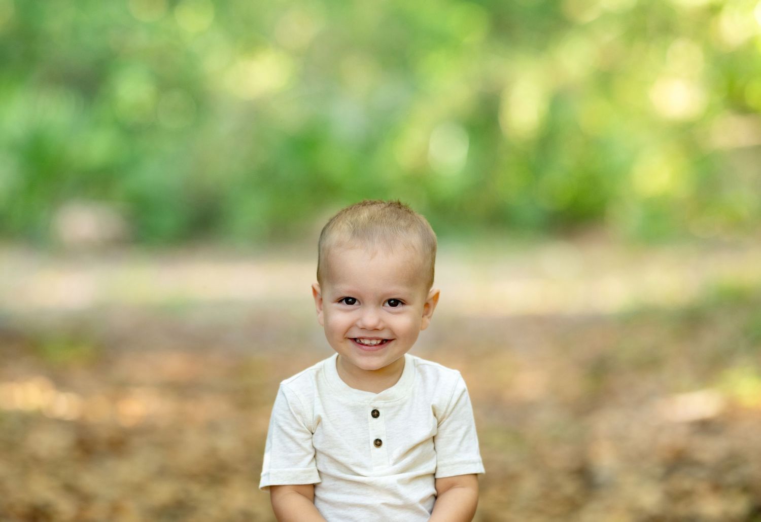 Smiling toddler boy in white shirt outdoors with green bokeh background.