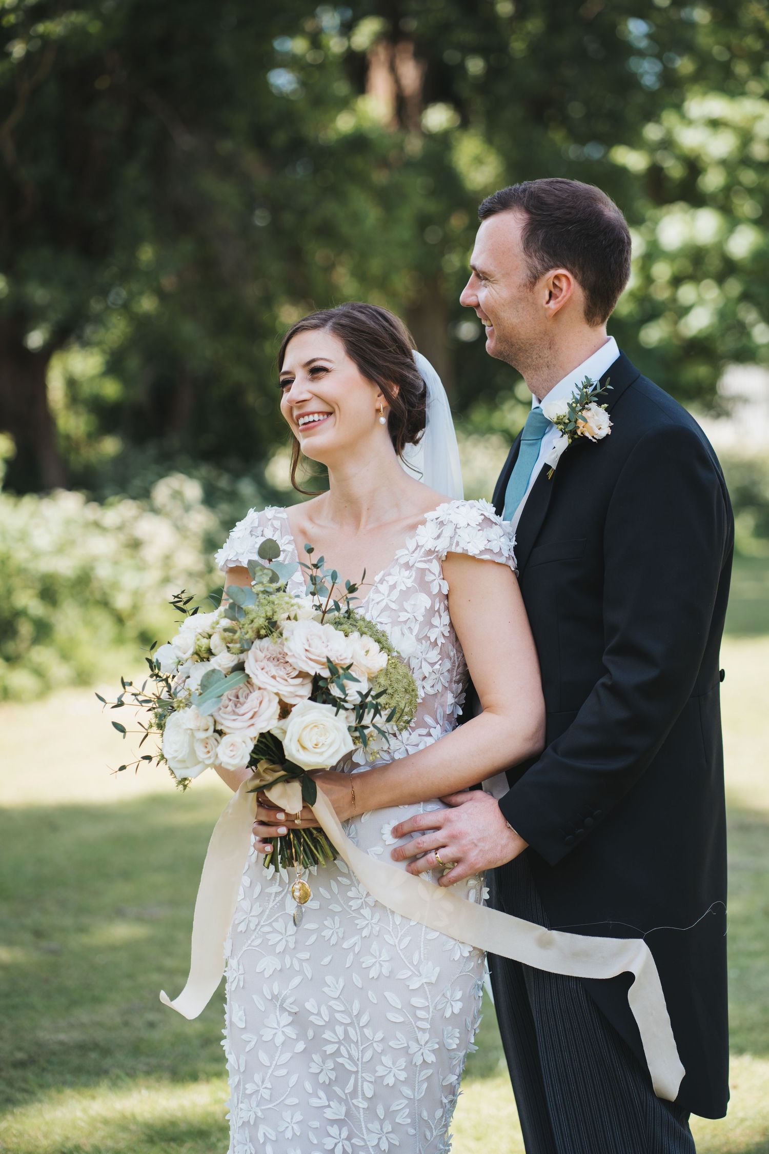 Bride in floral gown holds bouquet while groom embraces her from behind in sunny garden.