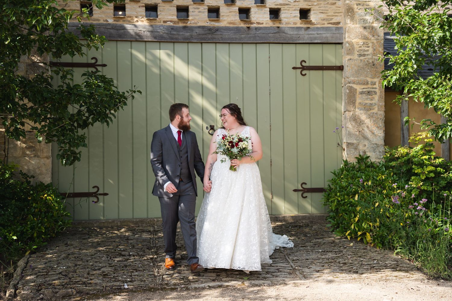 Bride and groom holding hands smiling at each other outside rustic stone barn with green doors on sunny wedding day at Oxleaze Barn Venue