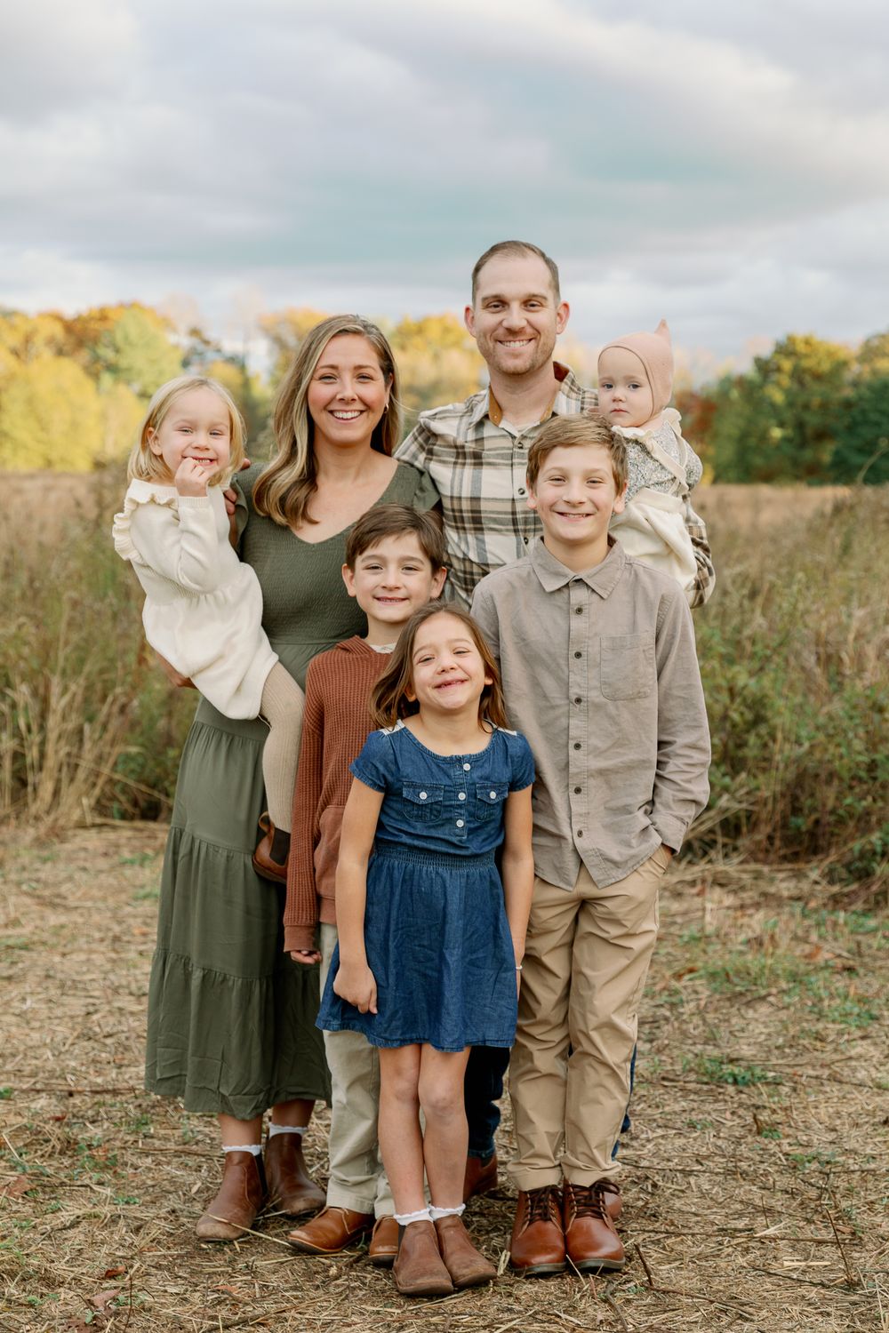 Family of seven posing outdoors in fall foliage, parents holding toddlers with children in coordinated autumn outfits.