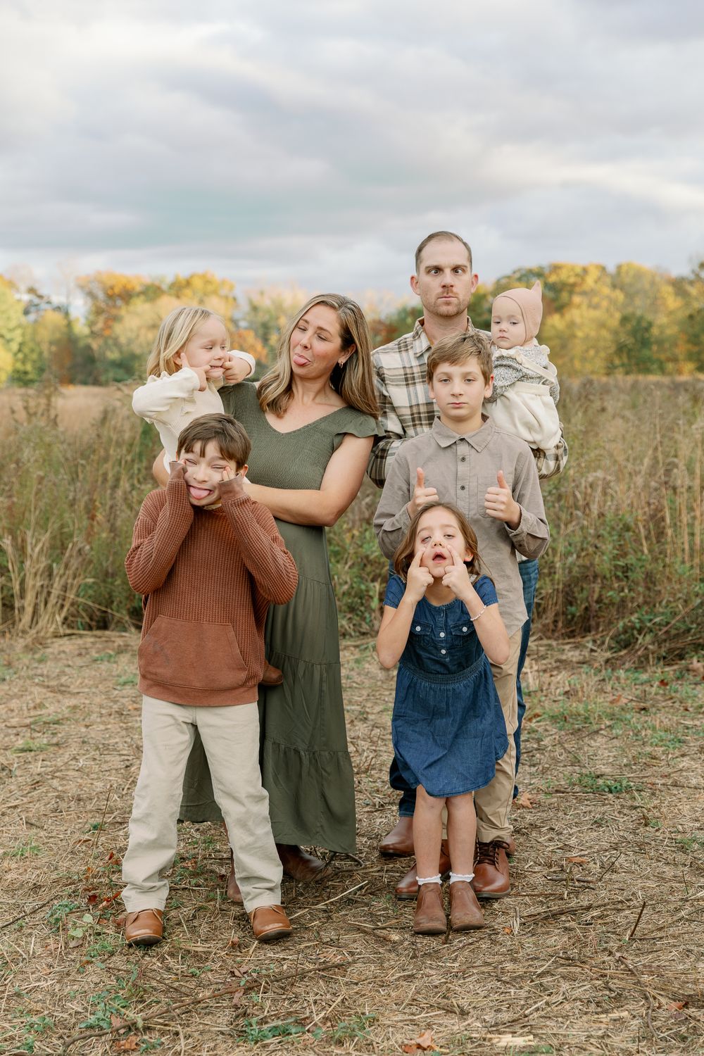 Family of six posing outdoors in a fall field, parents with four young children in autumn clothing.