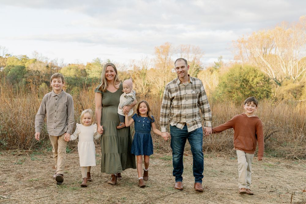 A joyful family of seven walks hand-in-hand through a golden autumn field, smiling together outdoors.