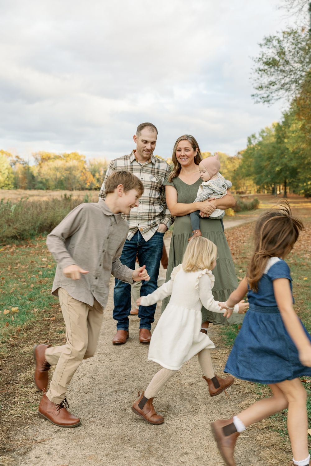 Family of six enjoying a playful autumn walk on a dirt path, parents smiling as children dance around them.