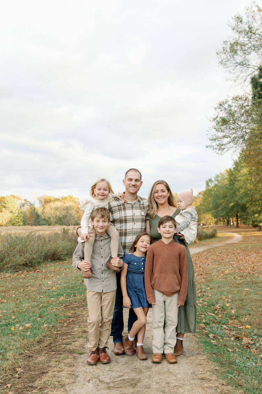 Happy family of six posing together on a dirt path with autumn trees in background.