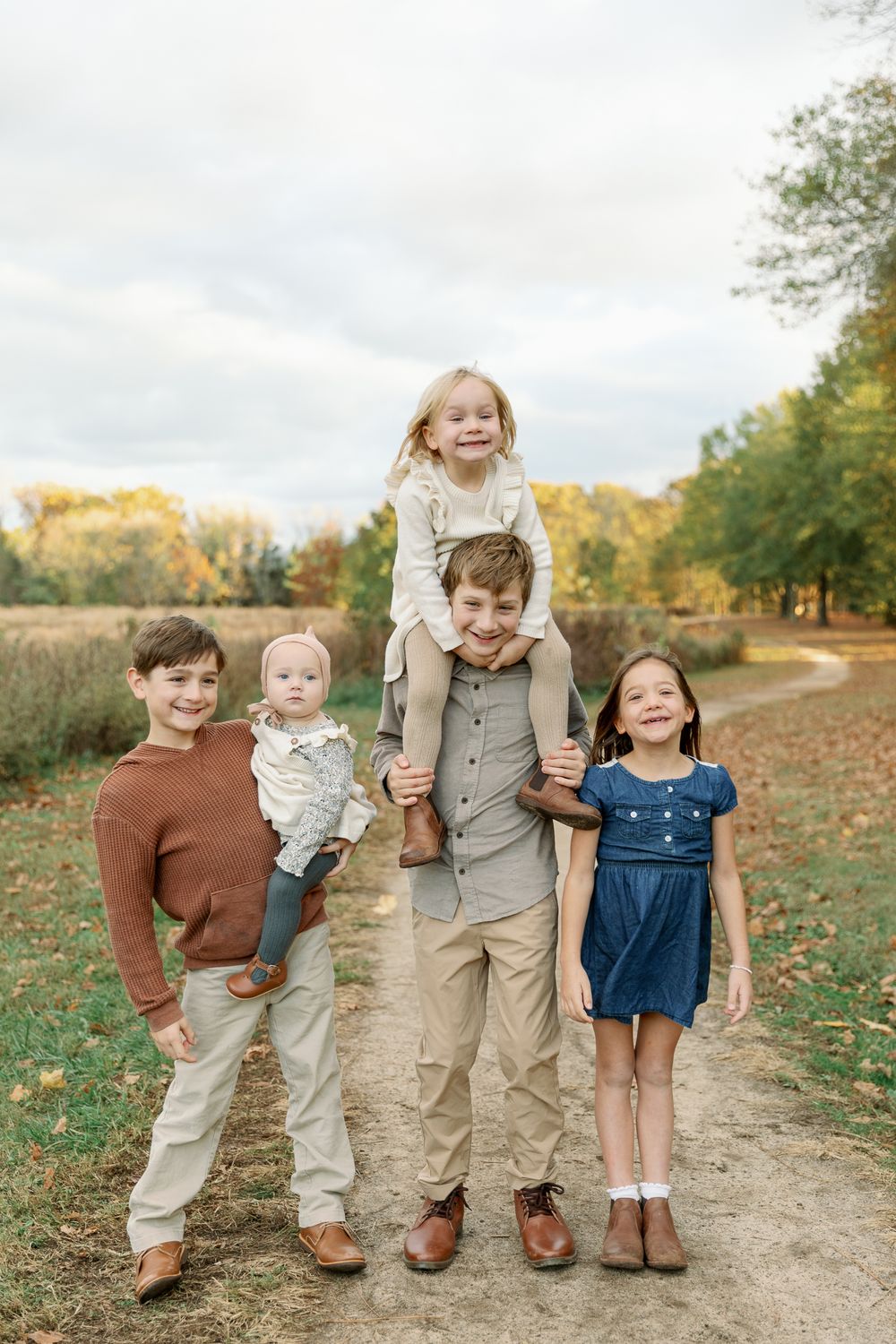 Five children pose on a dirt path surrounded by autumn trees, with the oldest boy giving a girl a piggyback ride.