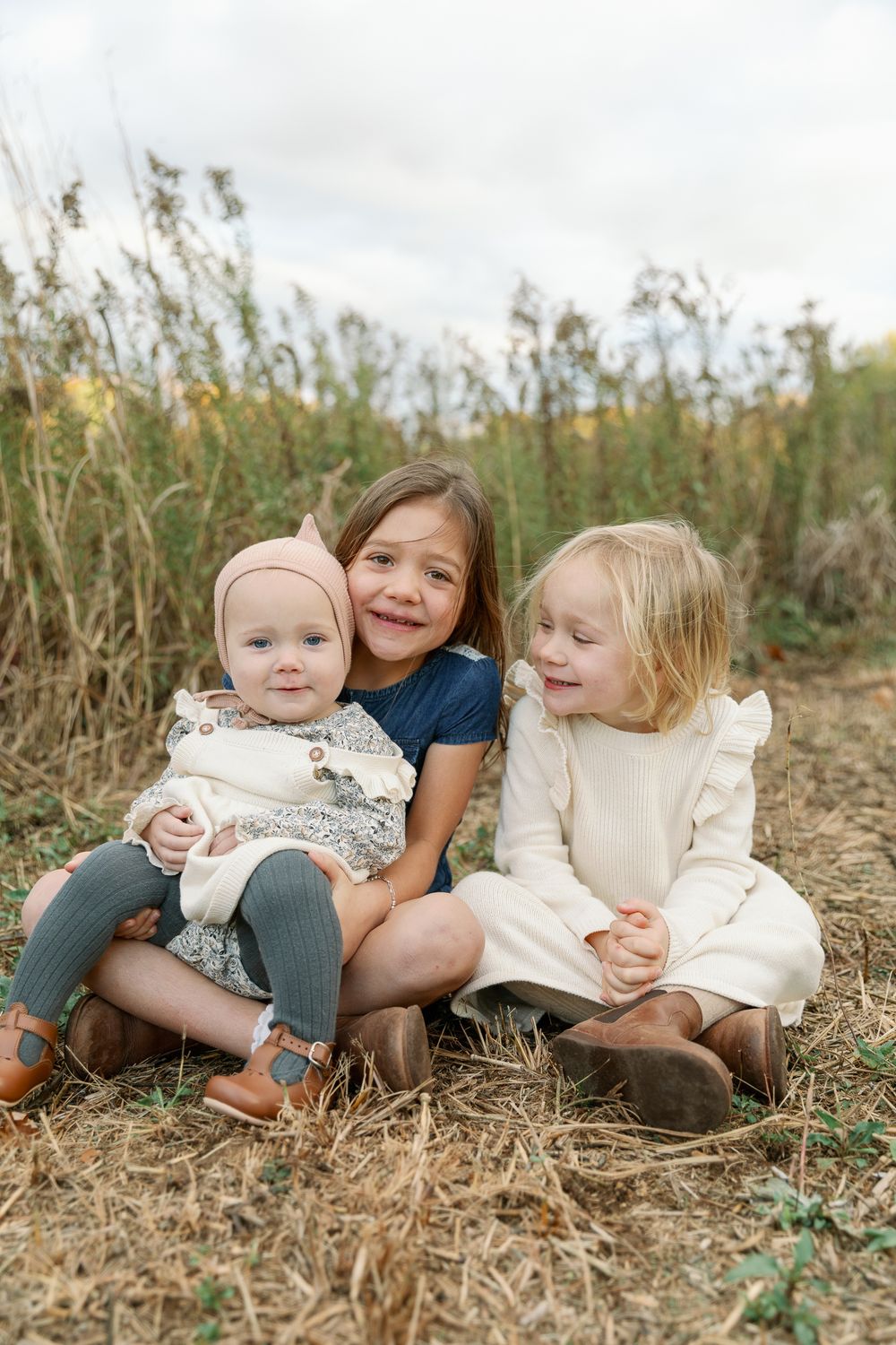 Three young sisters sitting together outdoors, oldest hugging baby sibling in fall setting.