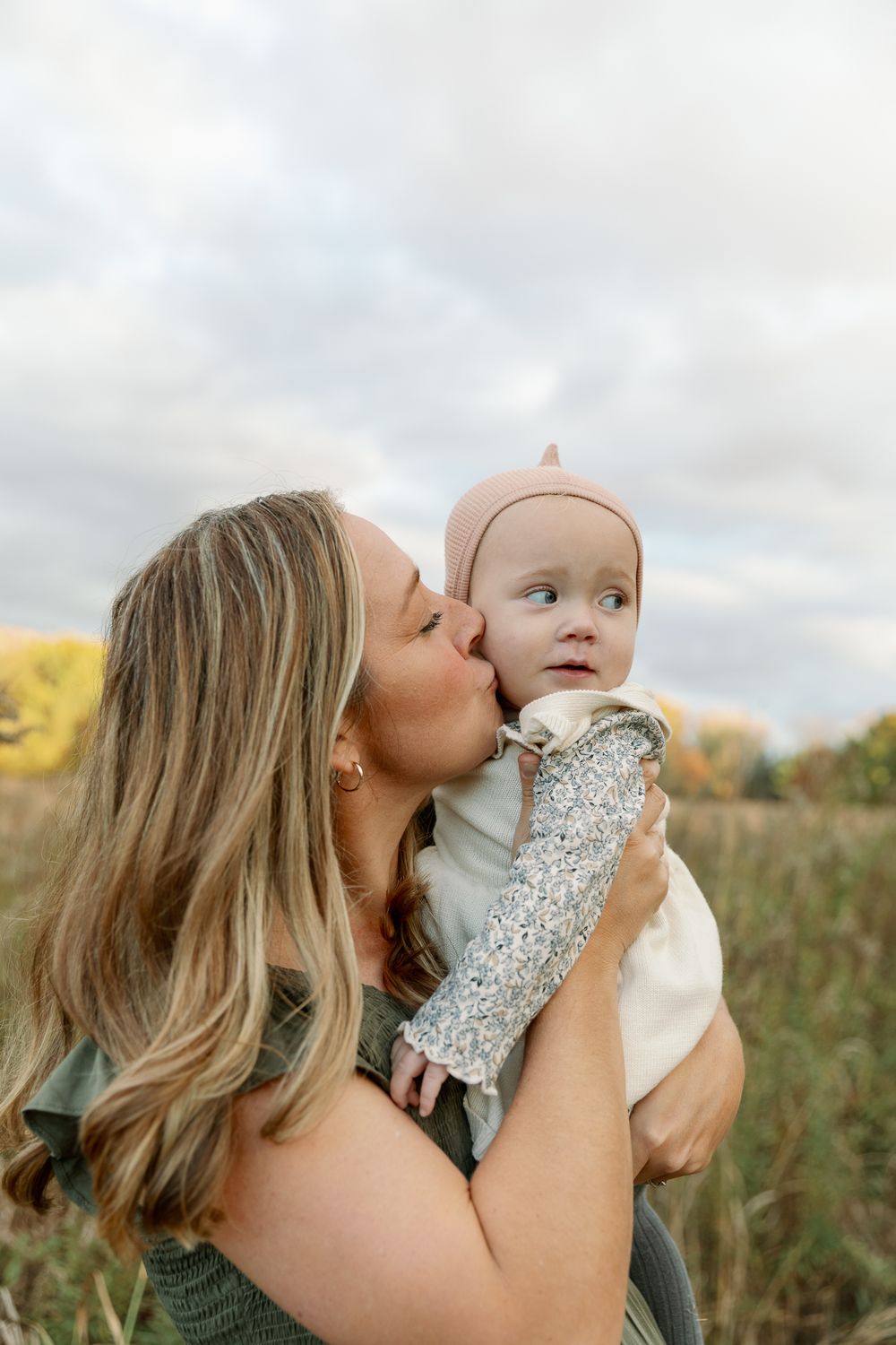 A mother tenderly kisses her baby wearing a pink dinosaur hat outdoors in a field with autumn trees.