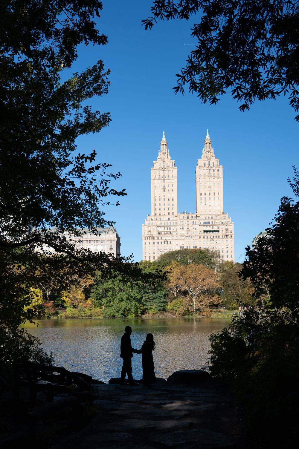 Couple silhouetted holding hands in Central Park with The San Remo twin towers and lake in background on clear day.