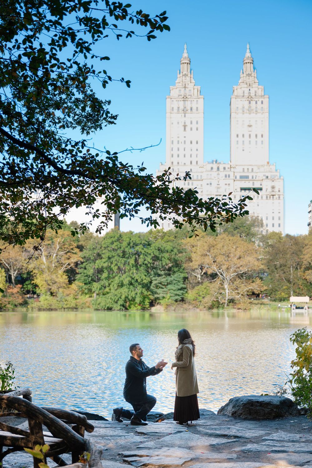 Man proposes to woman on one knee by a lake in Central Park, NYC, with the San Remo twin towers in the background.