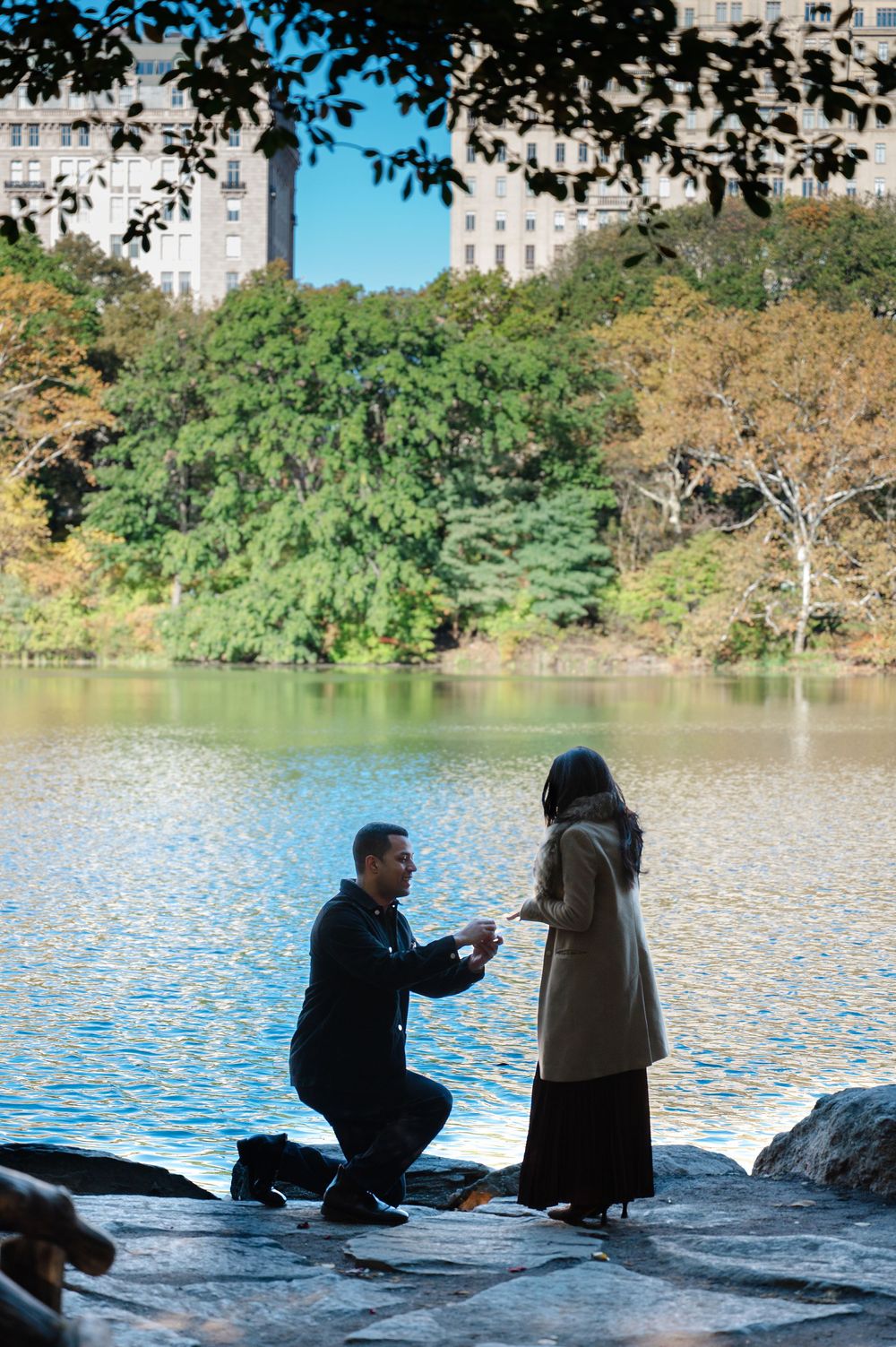 Man proposing on one knee by a lake in Central Park, New York, with trees and buildings in the background.