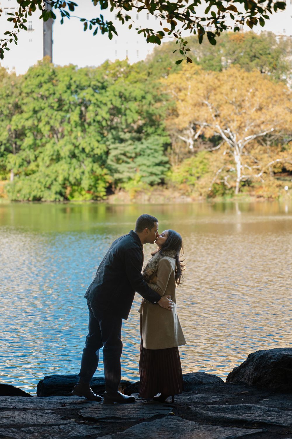 Couple sharing a romantic kiss by a serene lake surrounded by lush autumn trees in a peaceful park setting.