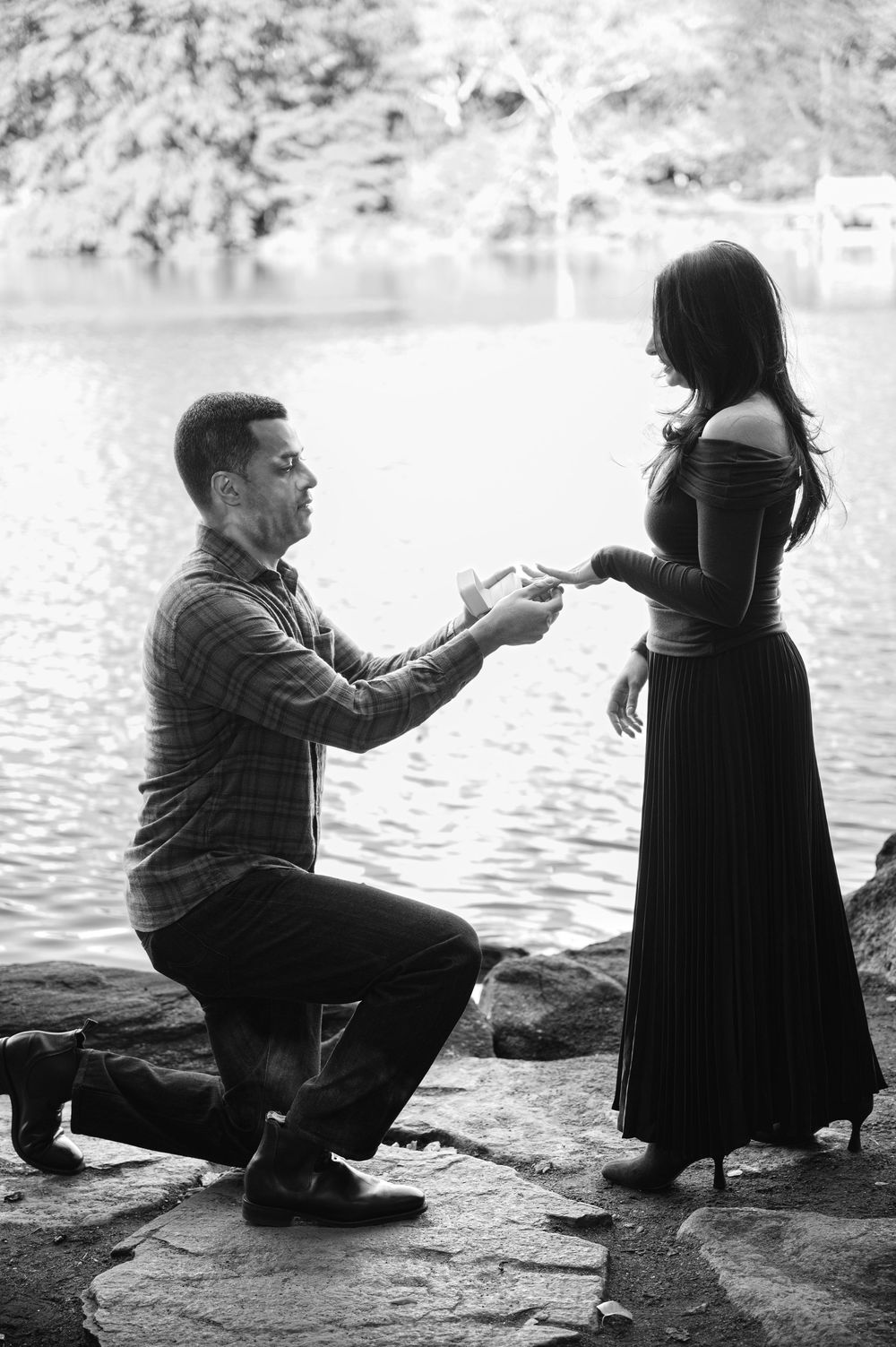 Man proposes on one knee to woman by a lake, holding a ring in a romantic black and white outdoor setting.