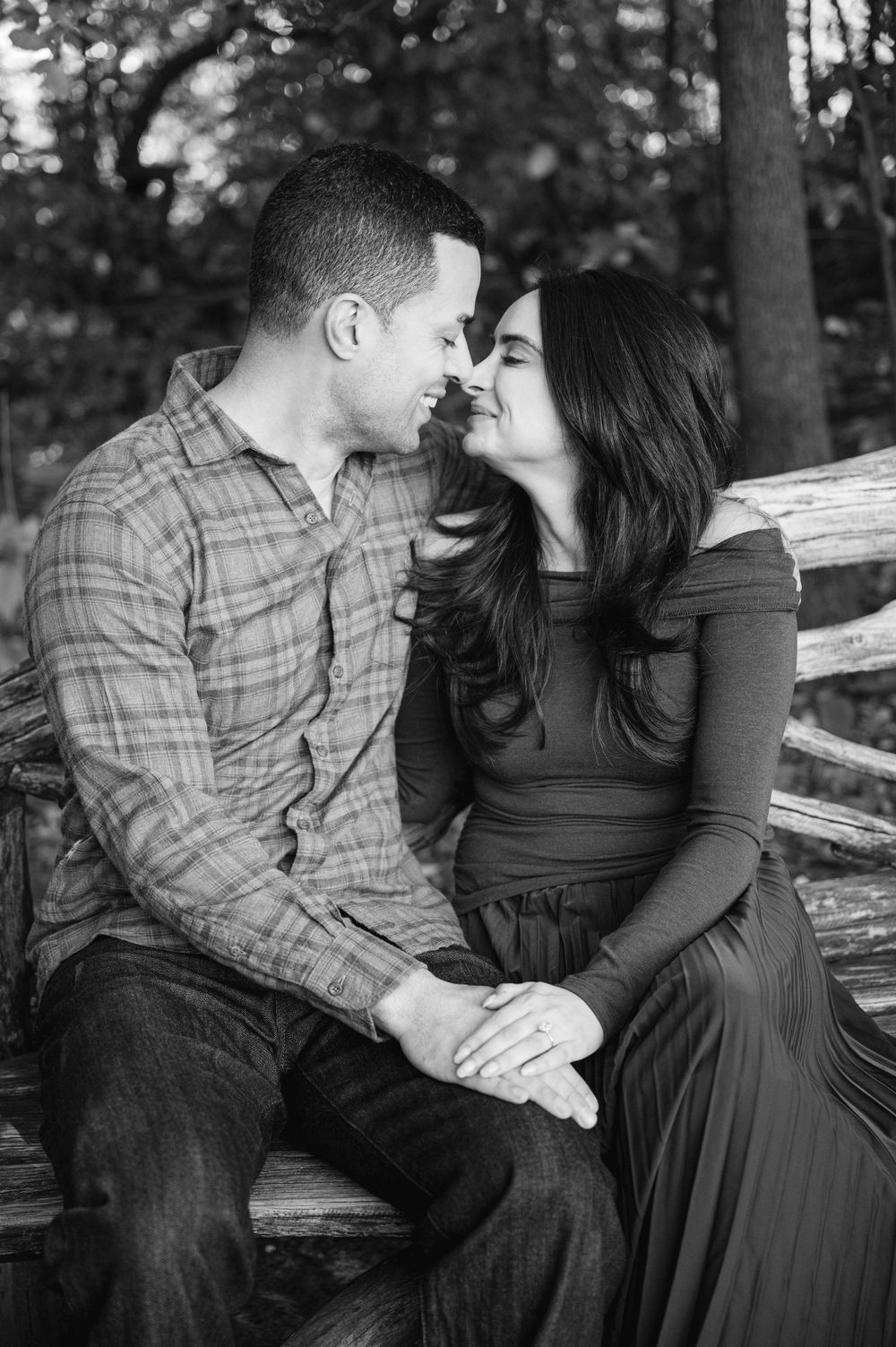 Black and white photo of a romantic couple sitting on a wooden bench, leaning in for a kiss outdoors.