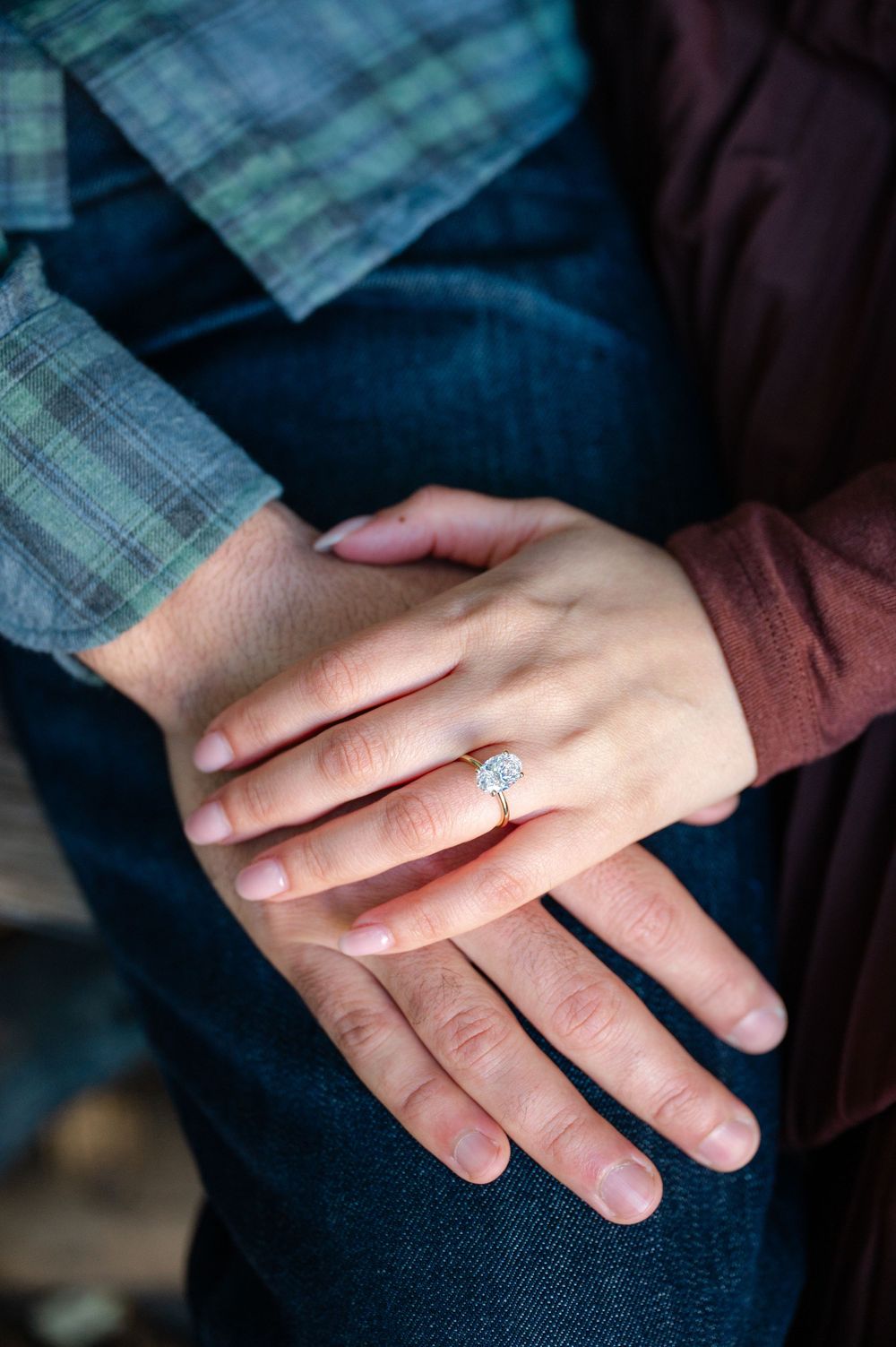 Close-up of couple holding hands showing diamond engagement ring, wearing plaid and burgundy outfits.