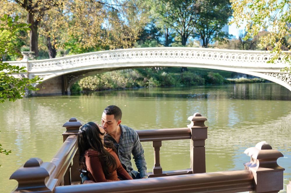 Couple kissing on a scenic bridge overlook at Central Park with Bow Bridge and a tranquil lake in the background.