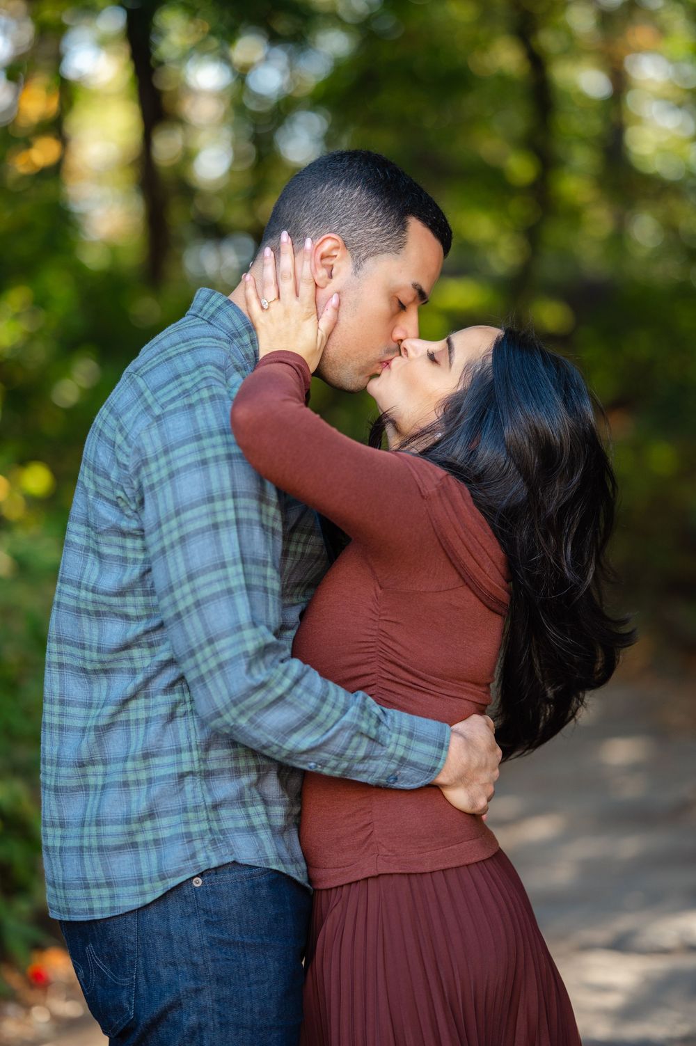Couple sharing a romantic kiss outdoors in a lush green park, man in plaid shirt embracing woman in rust dress.