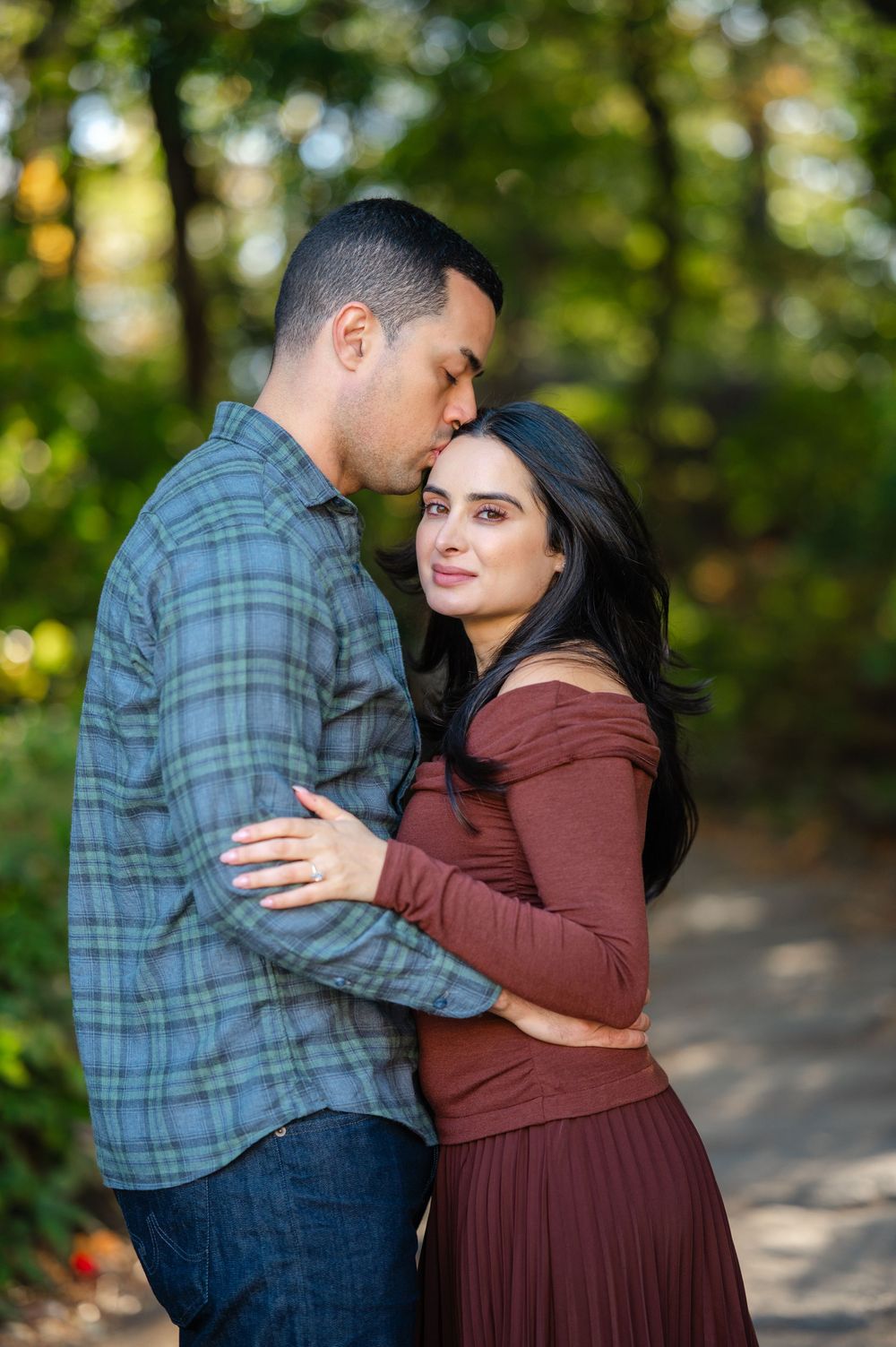 A couple embraces on a wooded path, man kissing woman's forehead, she wears a rust dress, he wears a plaid shirt.