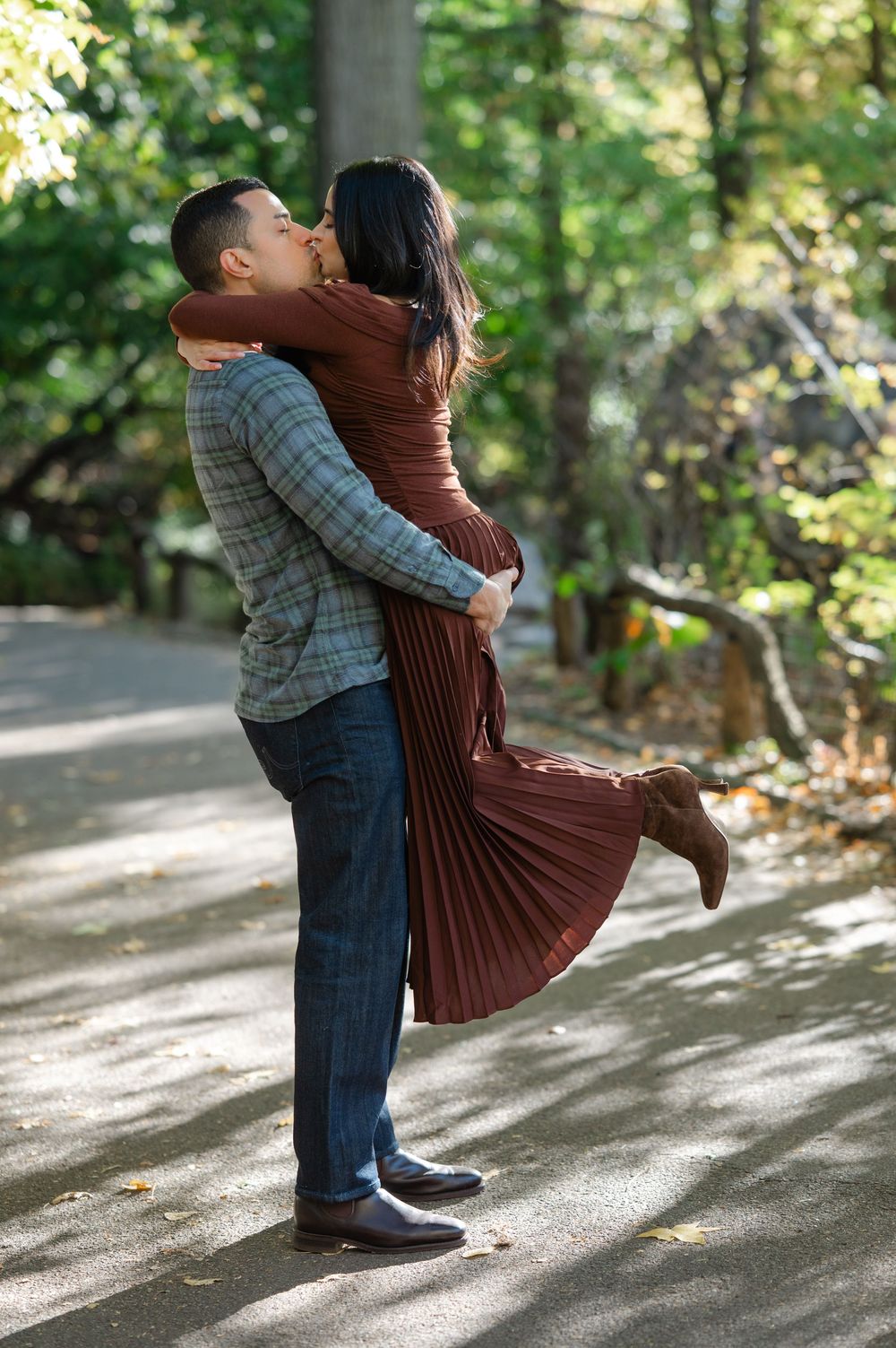 A couple shares a romantic kiss as the man lifts the woman in a sunlit park during fall.