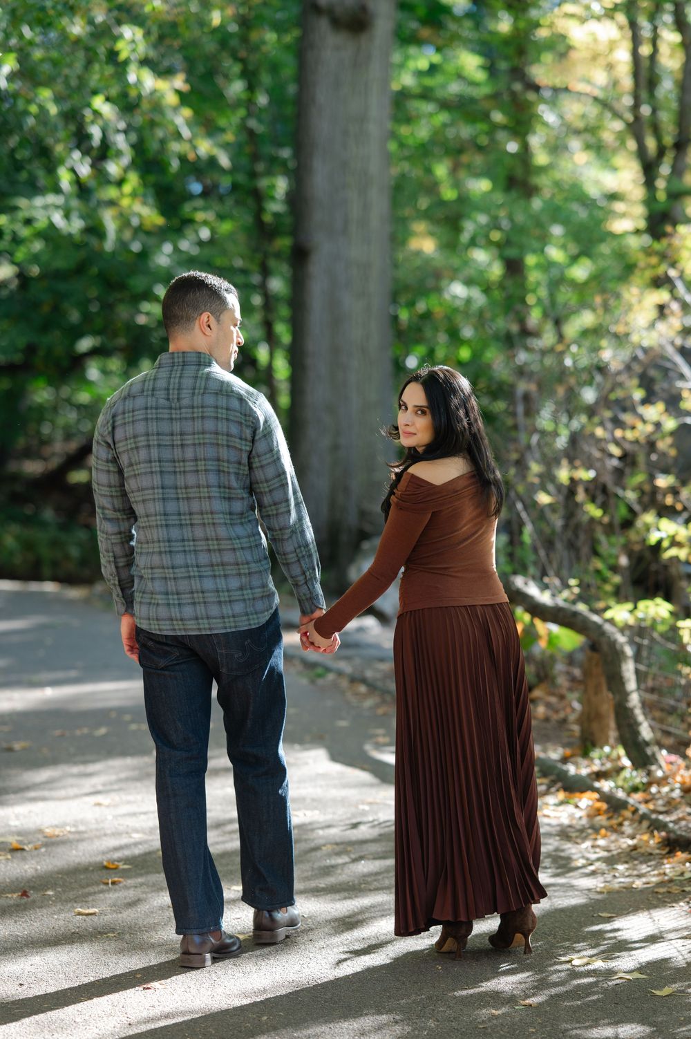 Couple holding hands on a sunlit park path in autumn, woman in brown dress looks back while man in plaid shirt faces away.
