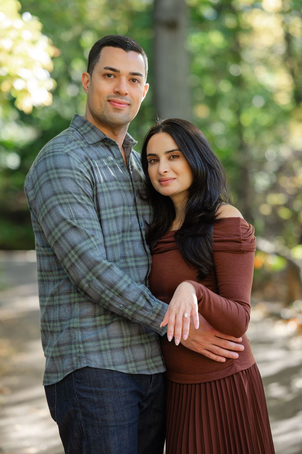 Couple posing together in a park, man in plaid shirt embracing woman in burgundy dress.